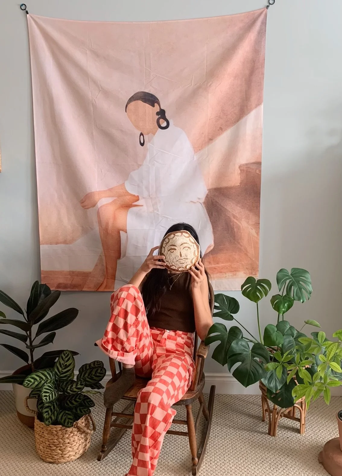 A woman sitting on a wooden chair holding a decorative face mask in front of her face, wearing checkered pants and a dark top, surrounded by green plants, with a large art piece of a woman’s illustration hanging on the wall behind her.