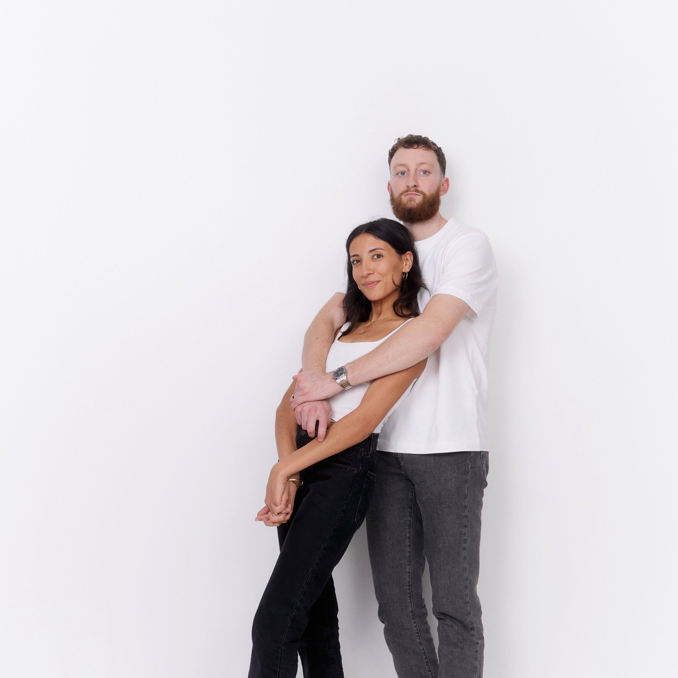 A man and a woman standing against a plain white wall, with the man standing behind the woman, both looking at the camera. The woman is smiling slightly, and the man has a neutral expression.