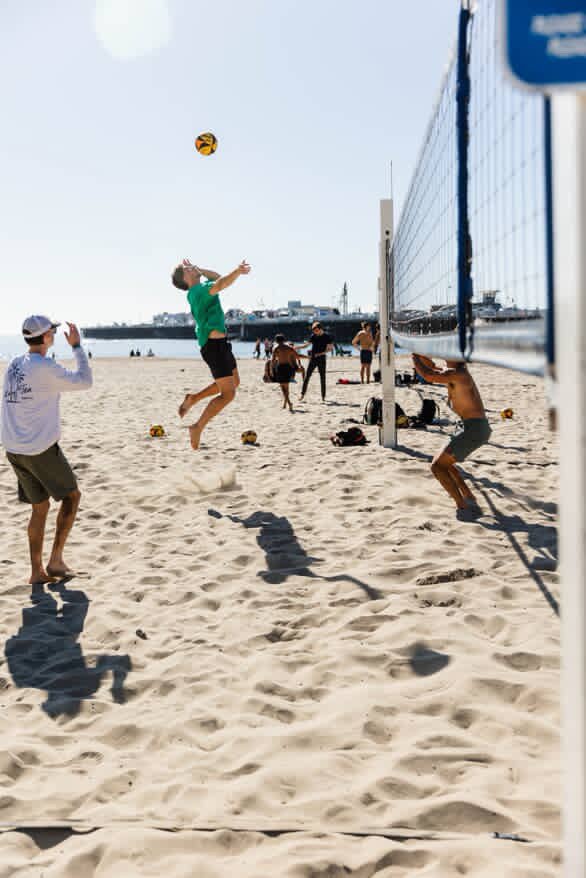 People playing beach volleyball on the sandy beach near water, with a few onlookers in the background.