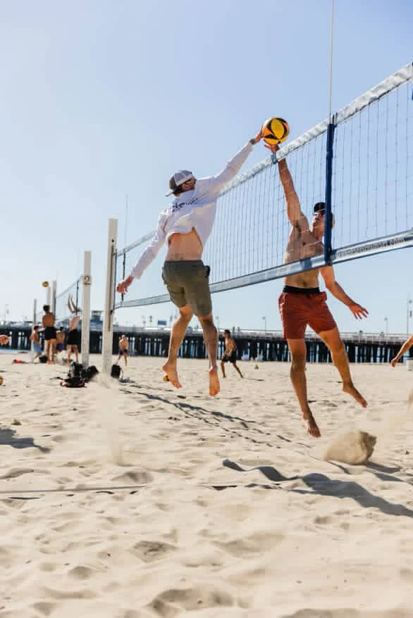 Two men playing beach volleyball near the ocean, one is jumping to hit the ball over the net while the other is jumping to block.