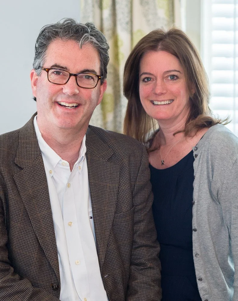 Smiling man and woman indoors, man in a blazer and glasses, woman in a black dress and cardigan