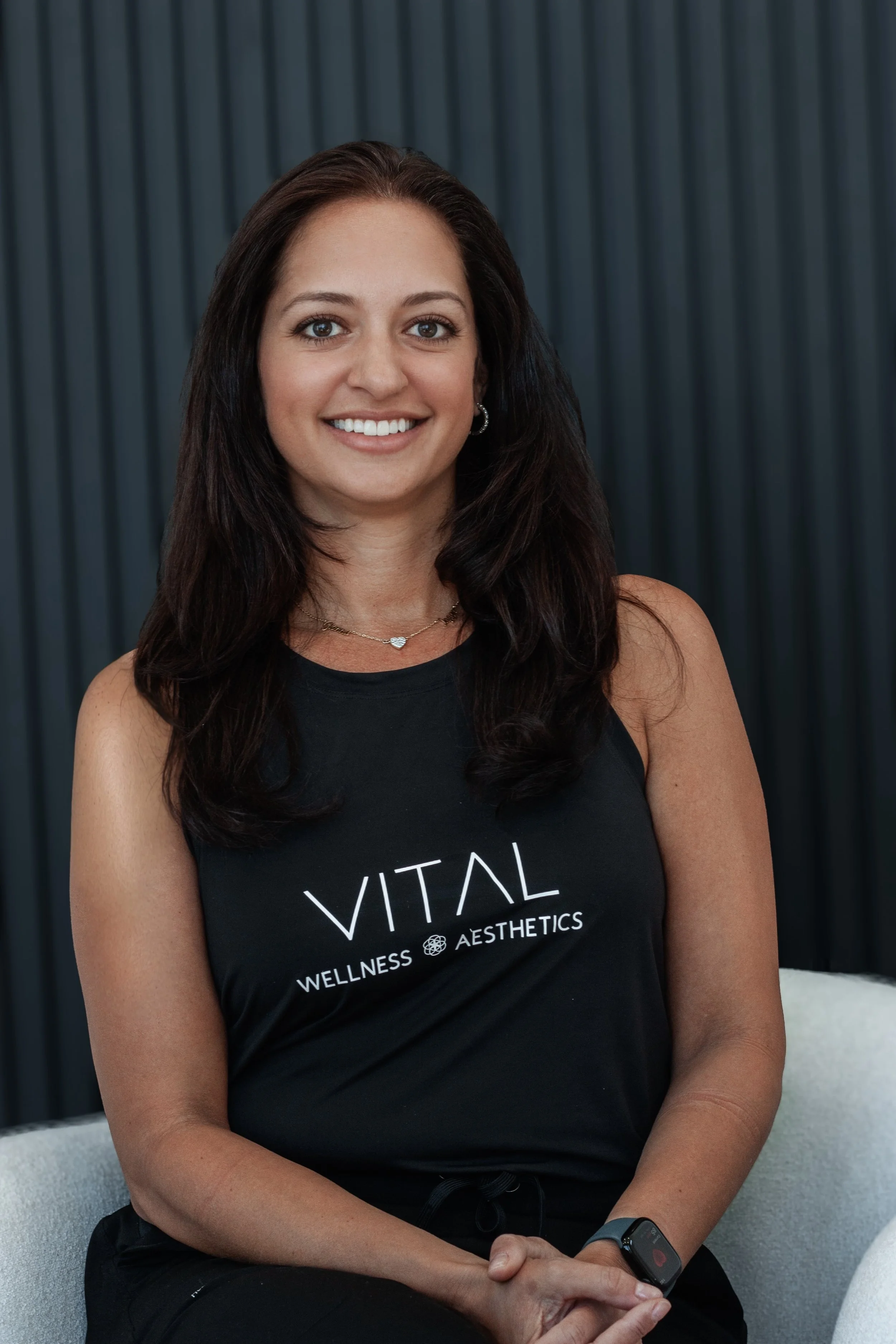 A woman wearing a black "Vital Wellness Aesthetics" tank top, sitting and smiling with a modern, dark striped background.