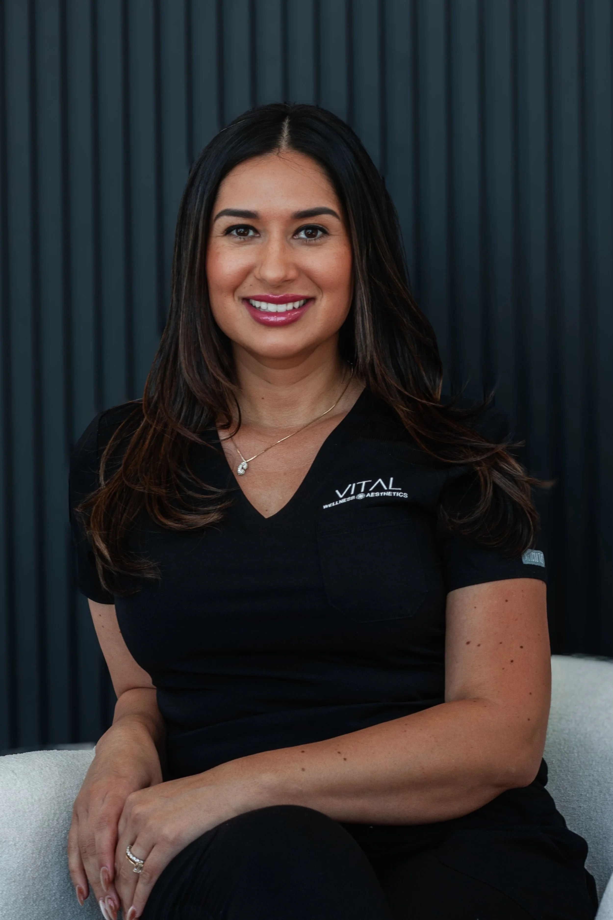 A woman with long dark hair, wearing a black top with a "VITAL Wellness & Aesthetics" logo, sitting in front of a dark, ribbed background. She is smiling, with her hands resting on her lap.
