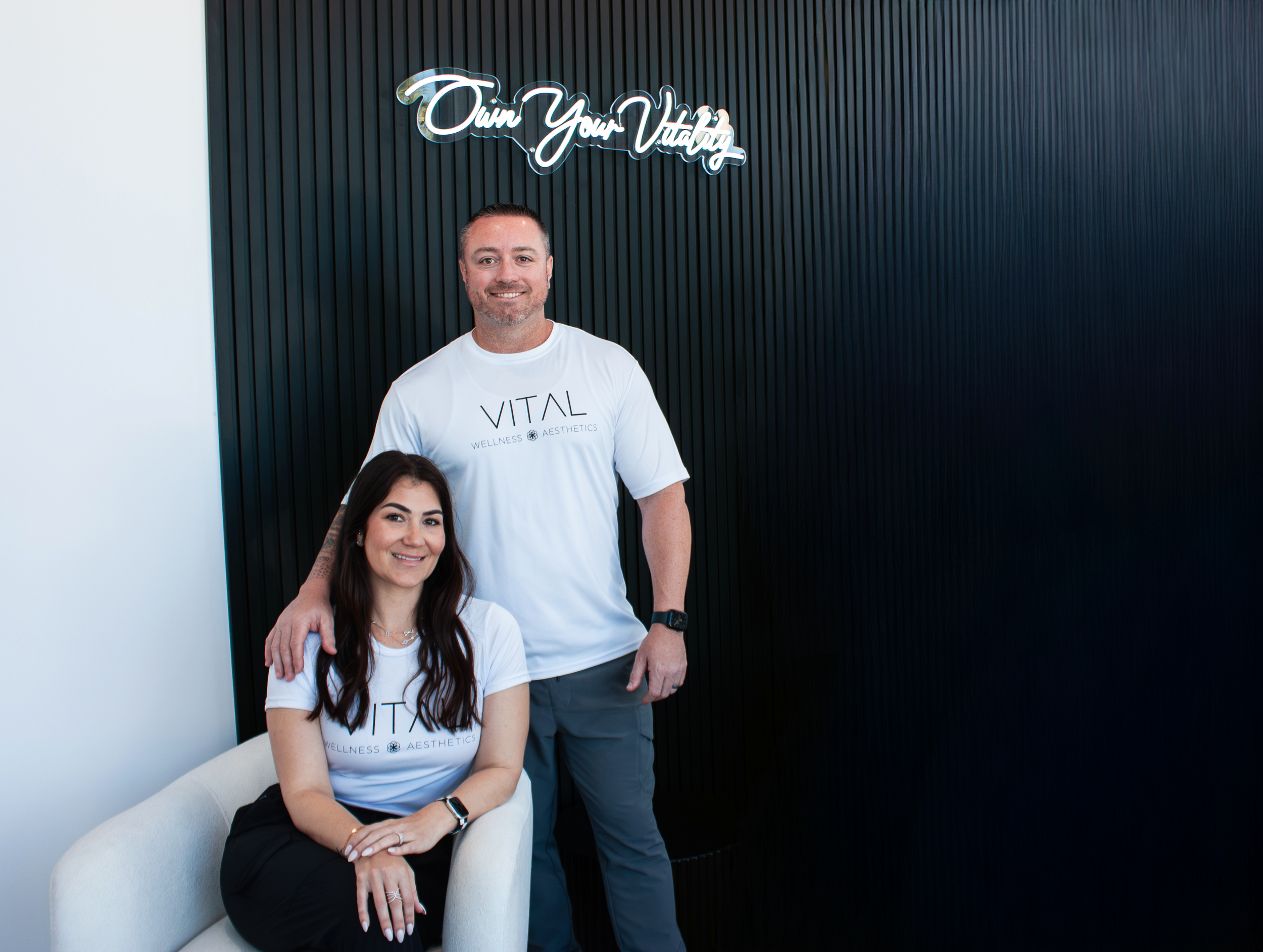 2 people wearing white "Vital Wellness & Aesthetics" shirts standing together in a room, with a "Own Your Vitality" sign on the wall behind them.