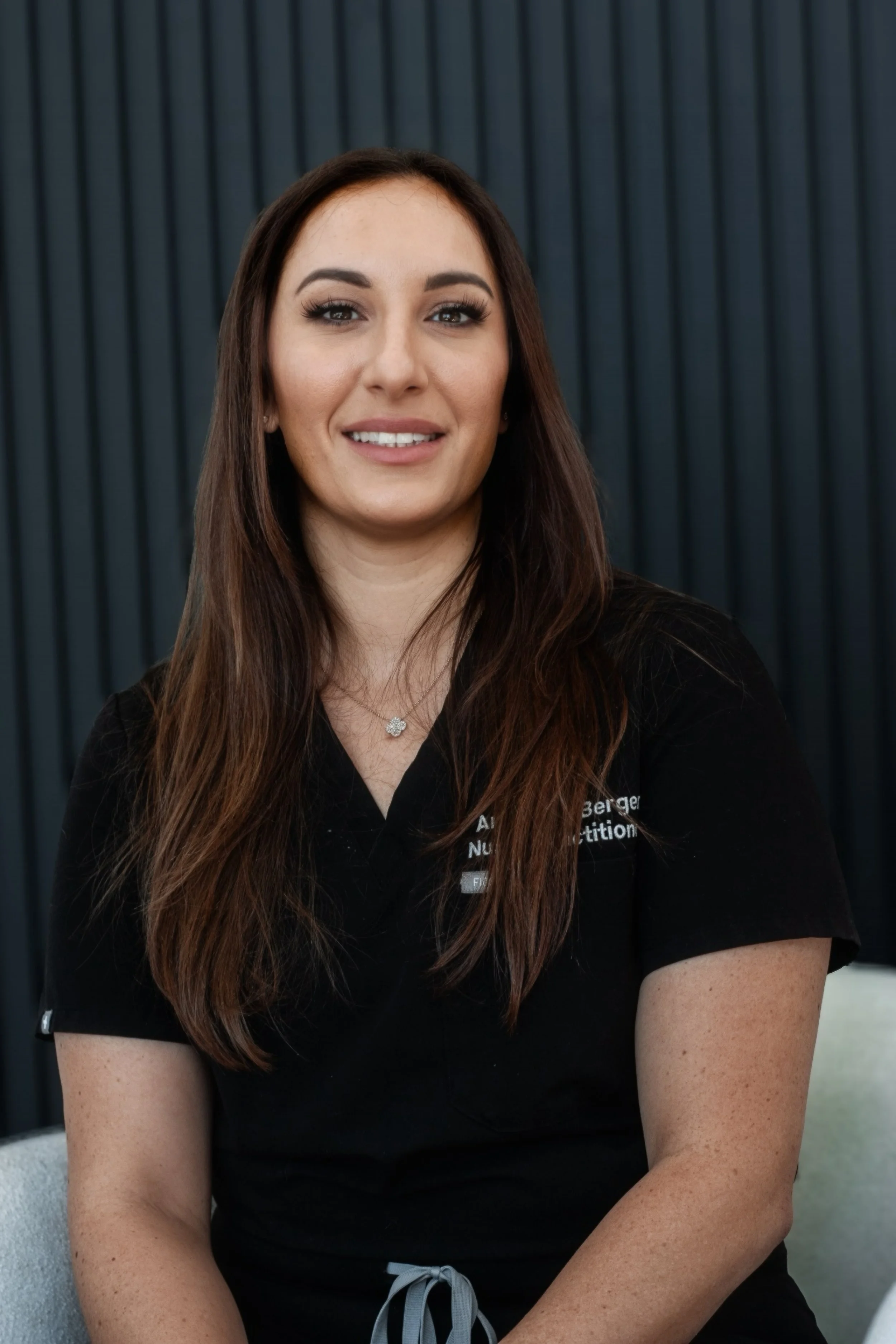 Smiling woman with long brown hair wearing a black uniform against a dark striped background.