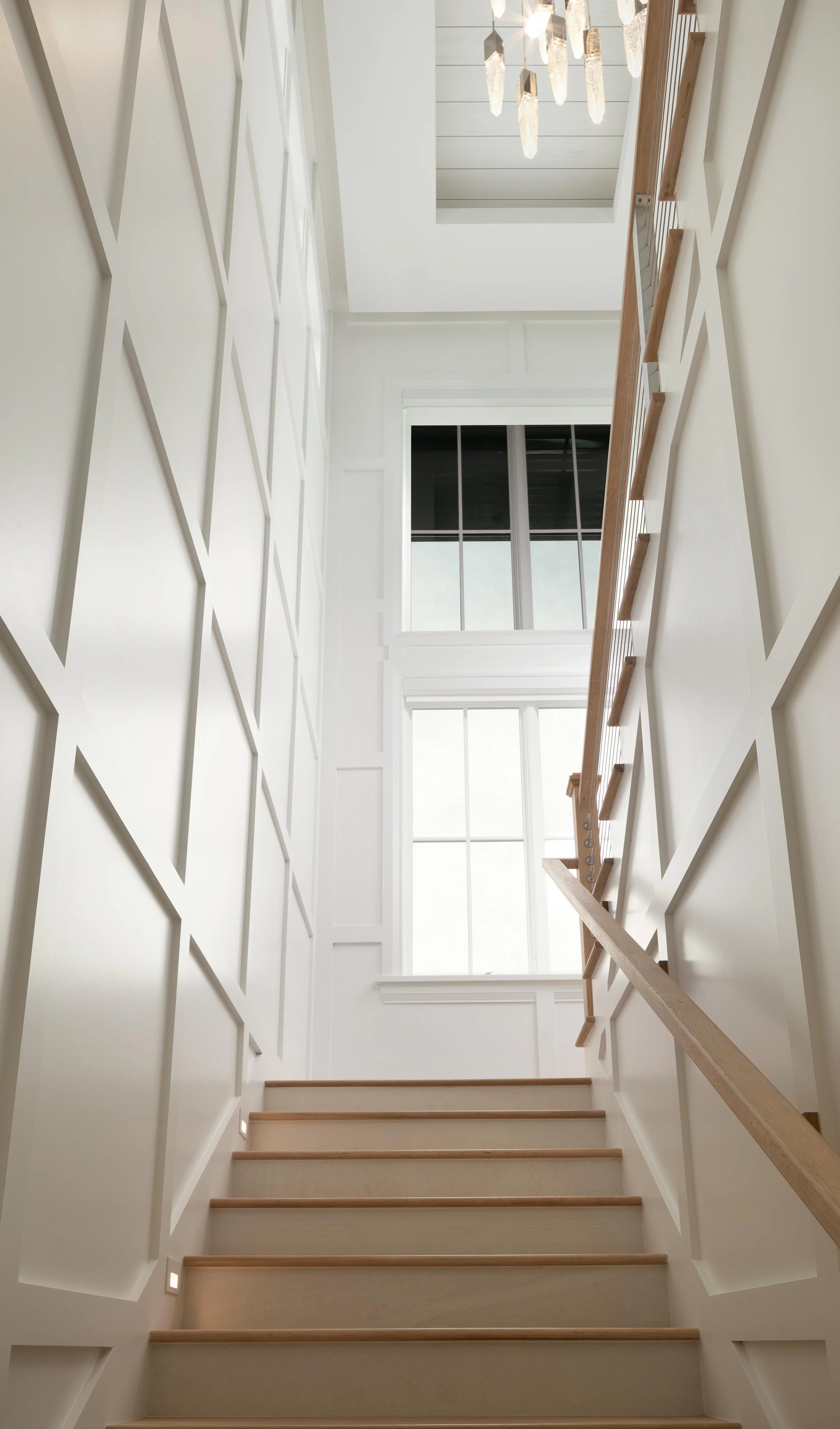 Modern staircase with wood railing and steps and recessed lighting, white paneled walls, and large windows. Elegant glass chandelier. Applied moulding geometric design by GLORIA BLACK for Naples Florida Talis Park New Construction Build home