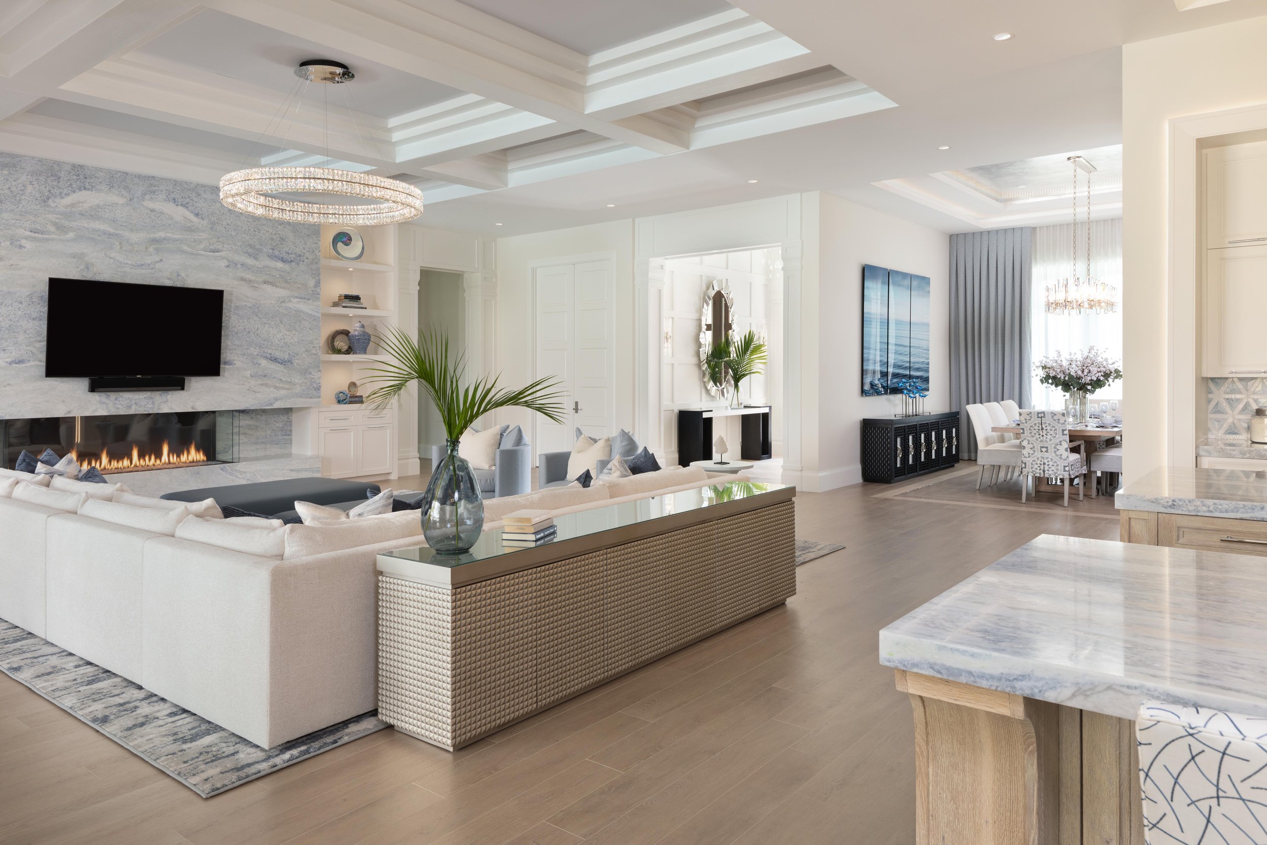 Modern living space with open floor plan featuring a large white sectional sofa, wall-mounted TV above a linear fireplace, intricate ceiling design, dining area with a table and upholstered chairs, and decorative plants. Light decor and spacious layo