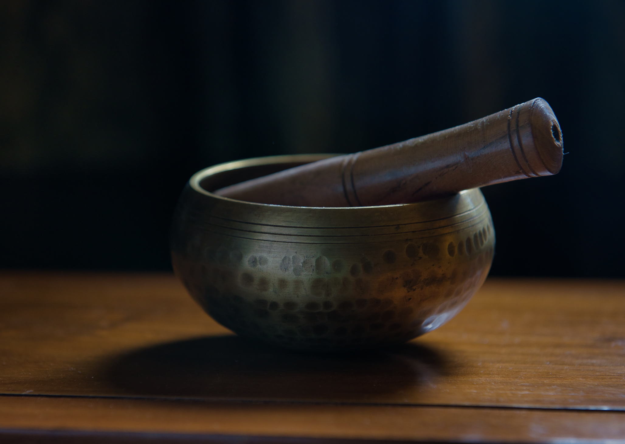 A traditional singing bowl with a wooden striker resting inside, placed on a wooden surface, with a dark background.