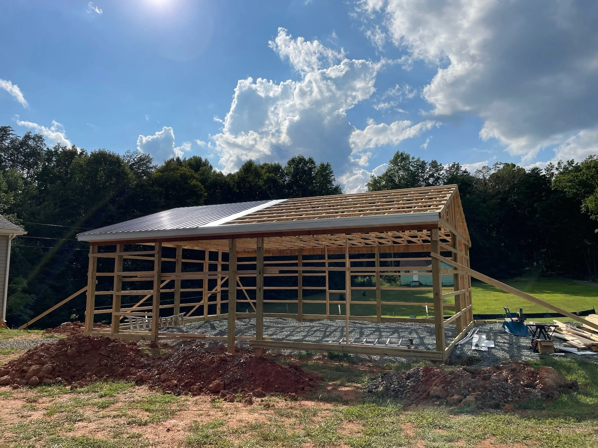 A construction site with a partially built wooden structure, a silver metal roof, surrounded by grass and trees under a partly cloudy sky.