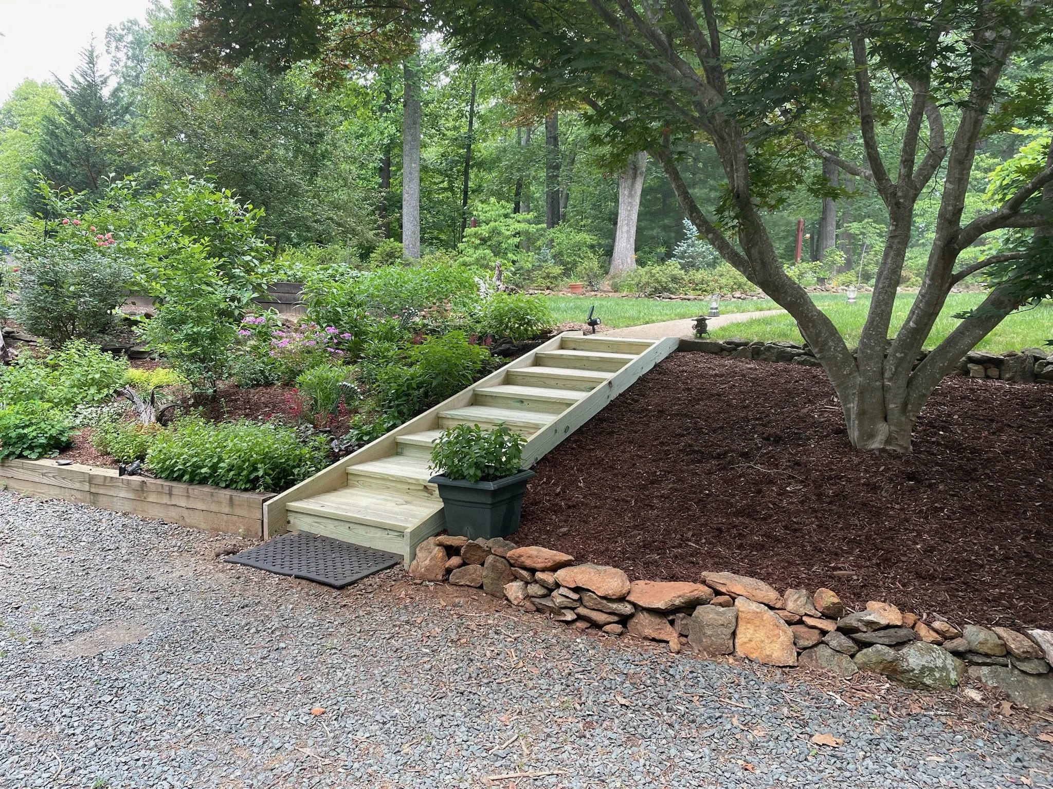 A backyard garden with a new wooden staircase leading from a gravel pathway to a grassy area surrounded by trees and various plants.