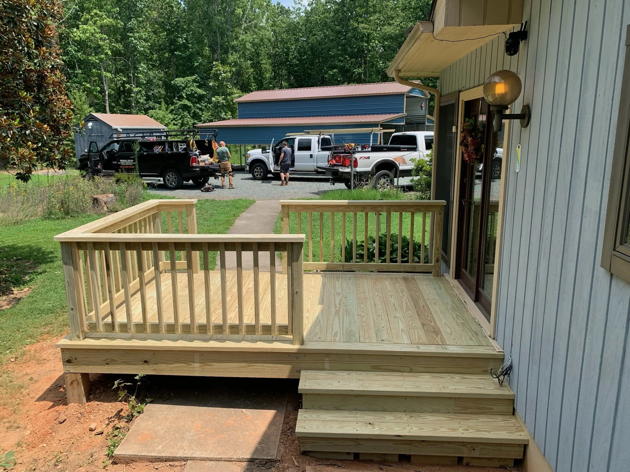 New wooden deck stairs and railing outside a house, with workers and trucks in the background.