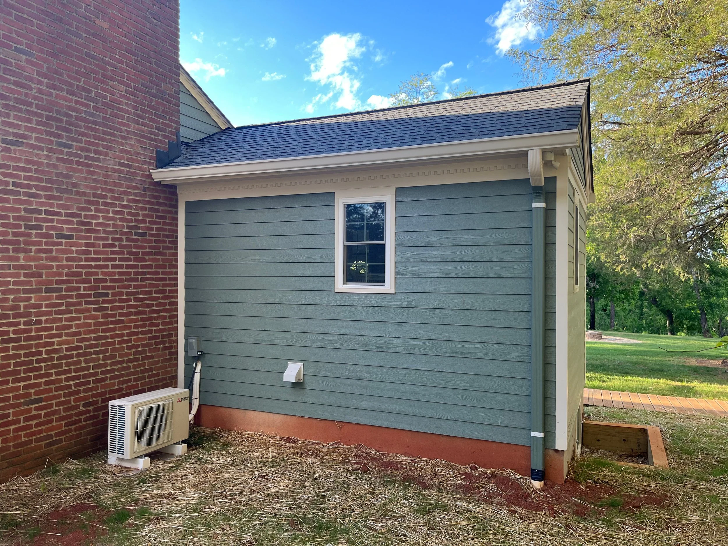 The photo shows the exterior corner of a house with a brick wall on the left and a blue siding wall on the right. A small window is on the blue wall, and an air conditioning unit is at the bottom left. The roof has dark shingles, and a white gutter runs along the edge. The area has grass and some trees in the background under a blue sky with some clouds.