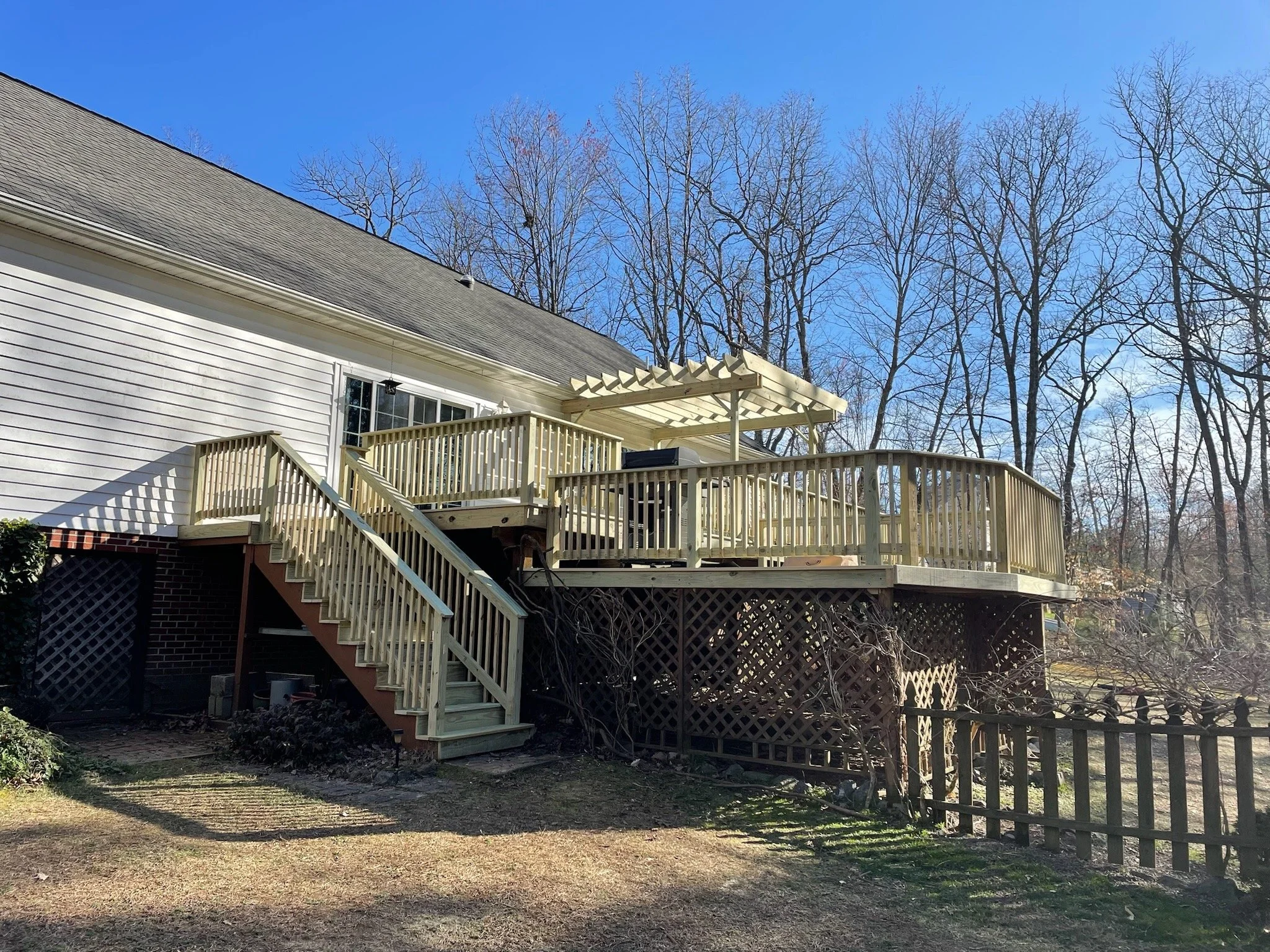 Backyard with a wooden deck, stairs, and an awning, house with white siding, and leafless trees in the background under a clear blue sky.