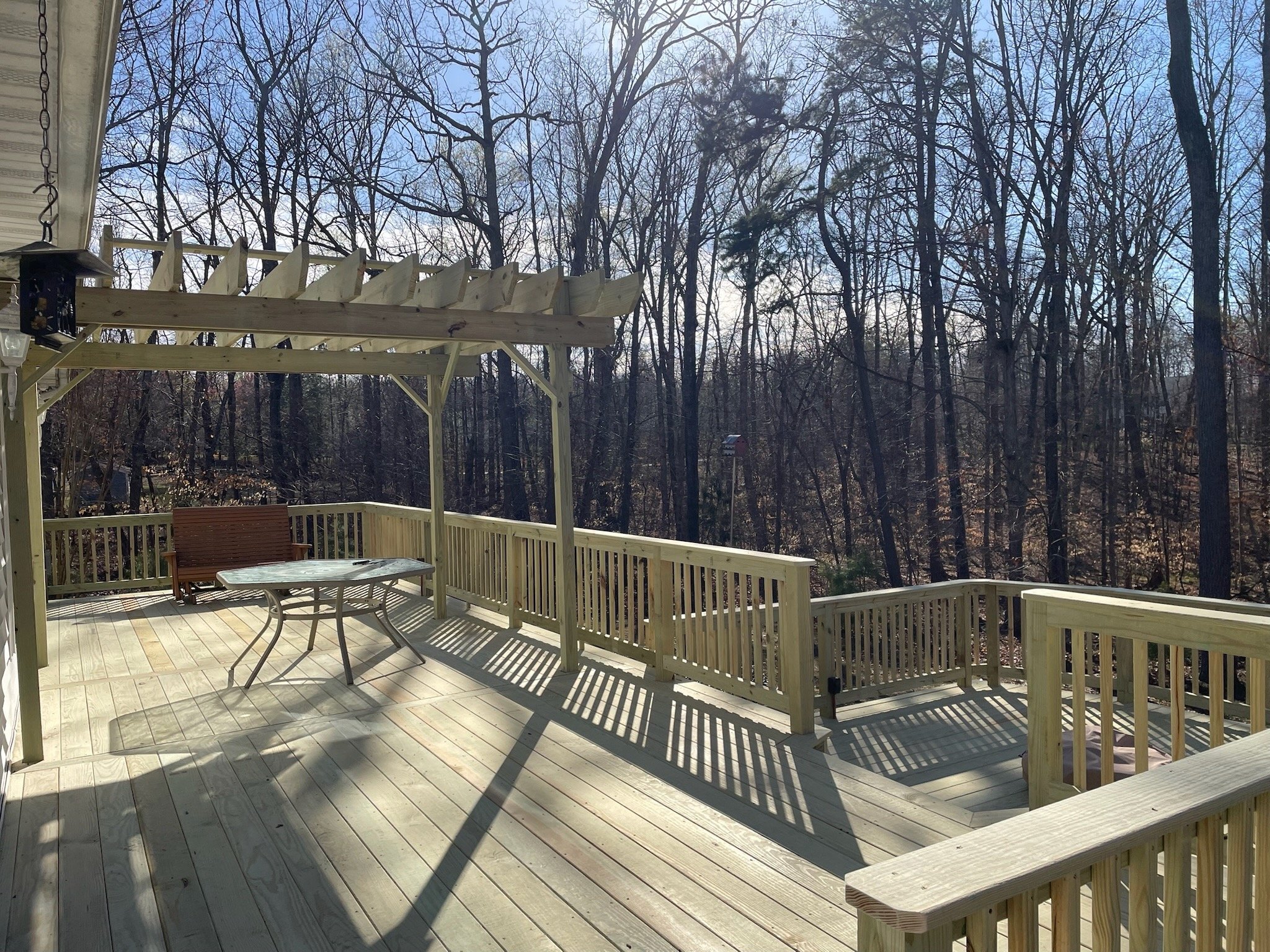A view of a backyard wooden deck with patio furniture and a wooden pergola, surrounded by leafless trees under a partly cloudy sky.