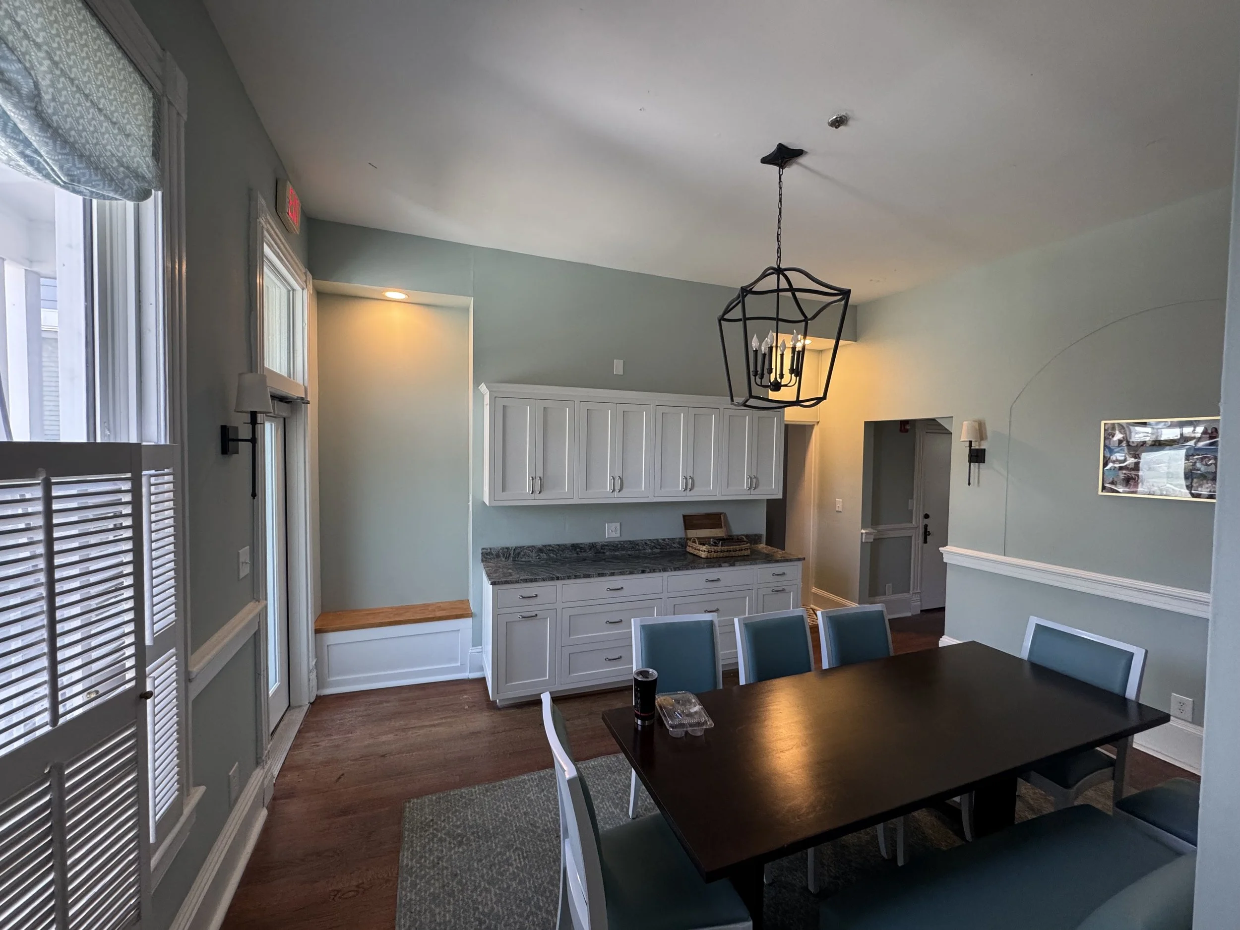 Interior of a dining area with a dark wooden table, blue chairs, white cabinets, a chandelier, and windows with blinds.