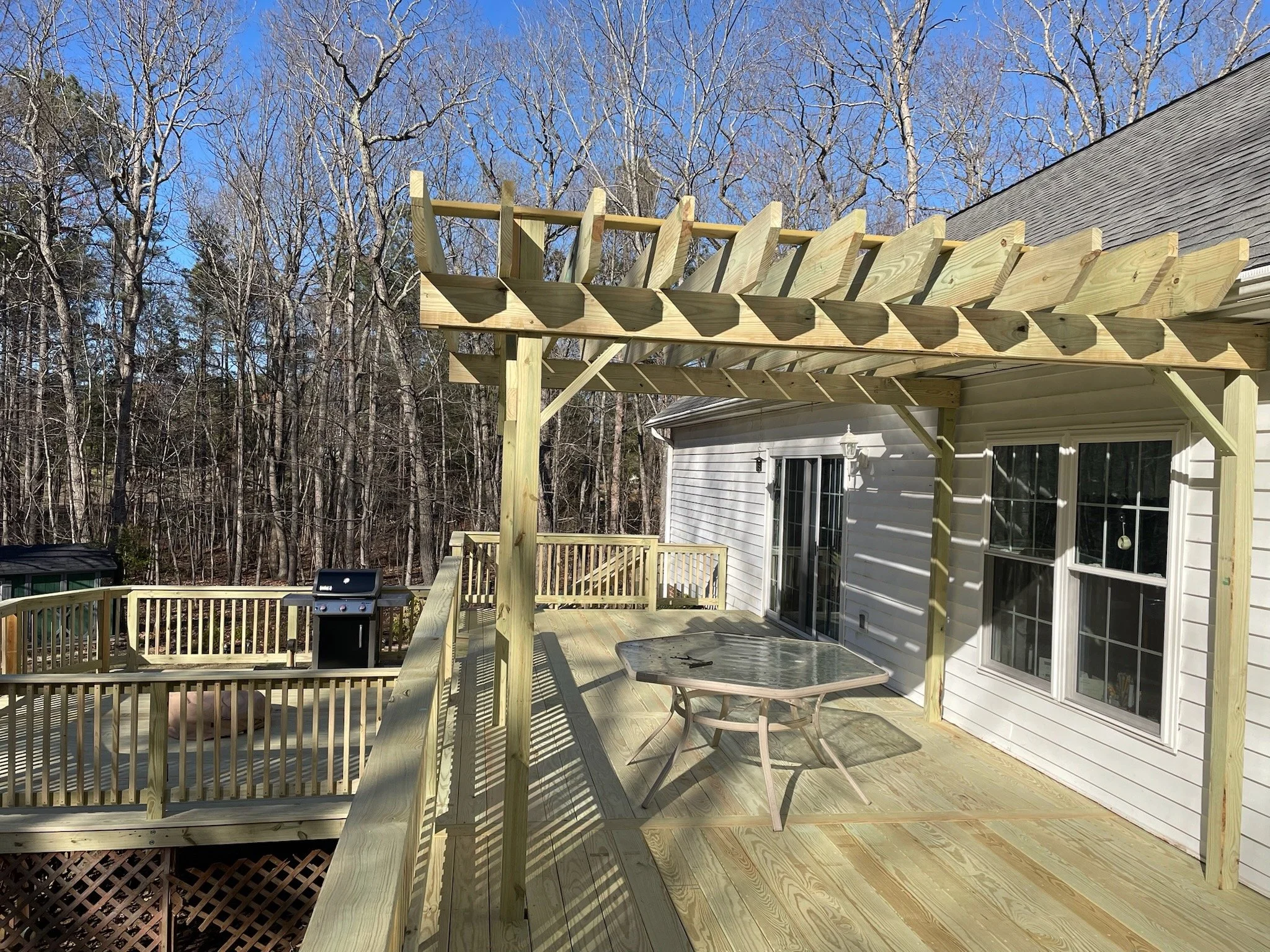 A wooden deck with a pergola structure under construction attached to a white house, with a surrounding wooded area and a clear blue sky.