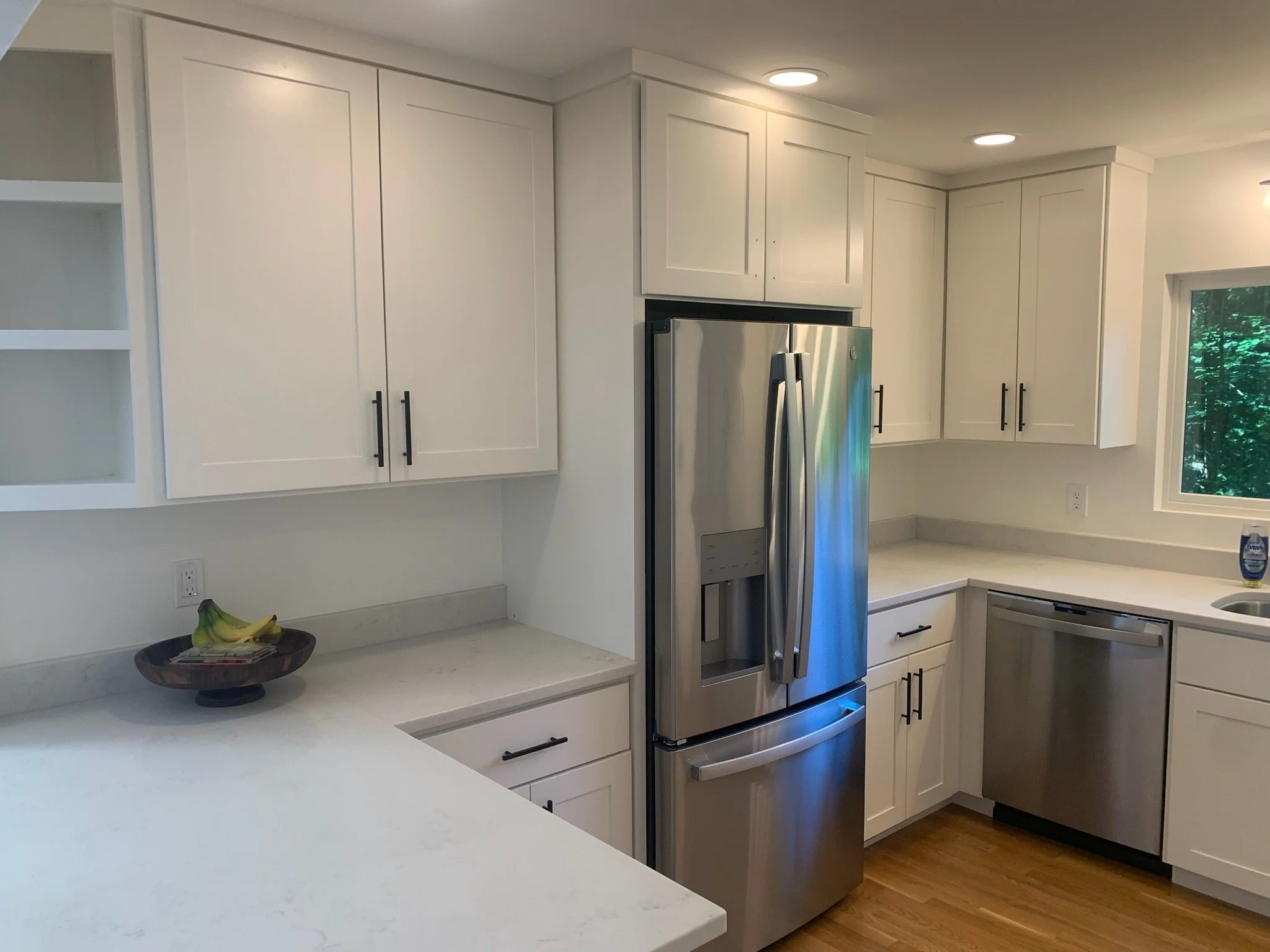 White modern kitchen with white cabinets, stainless steel fridge, and a window with greenery outside.