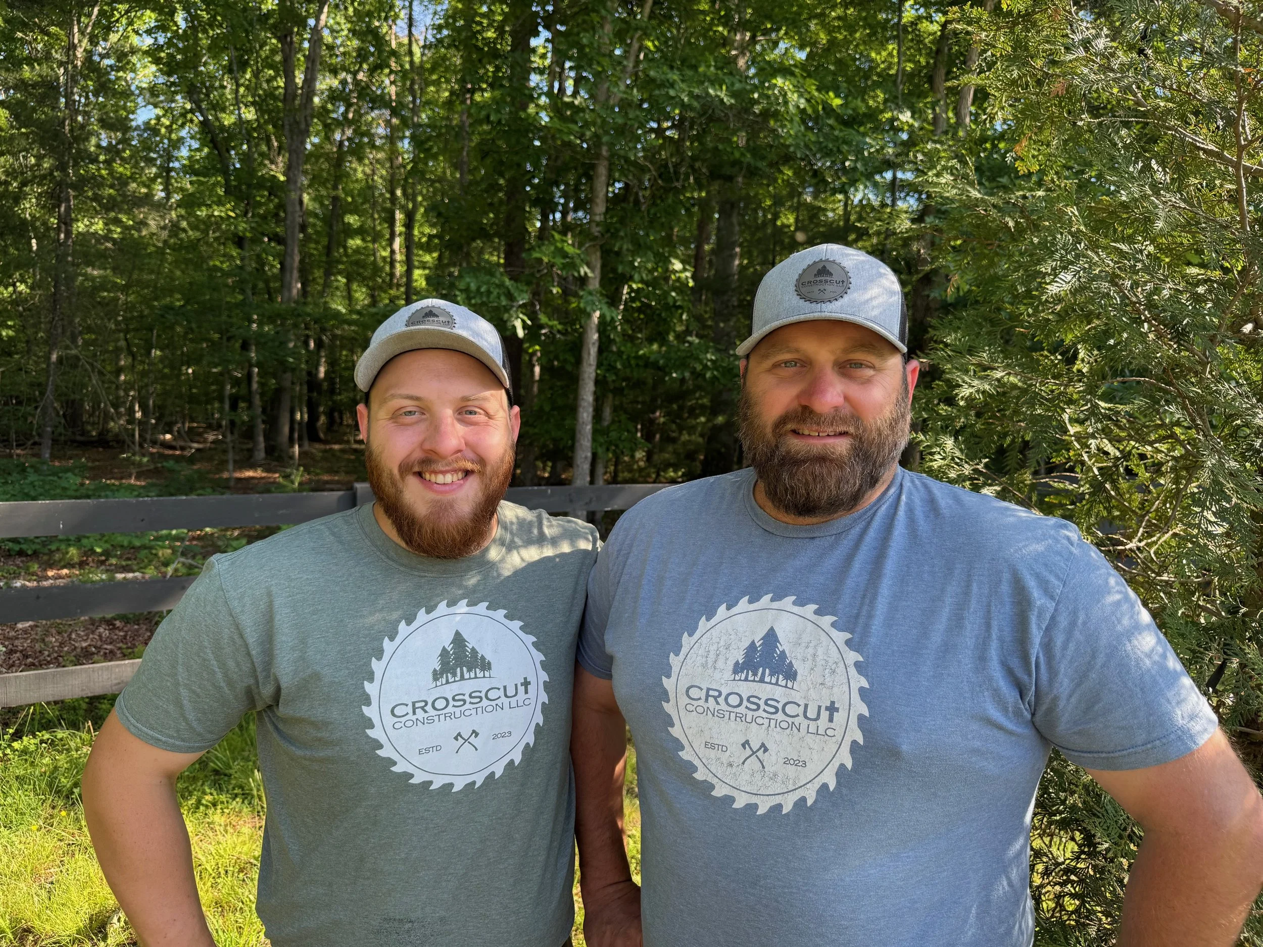 Two men wearing matching gray caps and gray t-shirts with a logo for Crosscut Construction LLC stand outdoors in front of a wooded area, smiling for the camera.