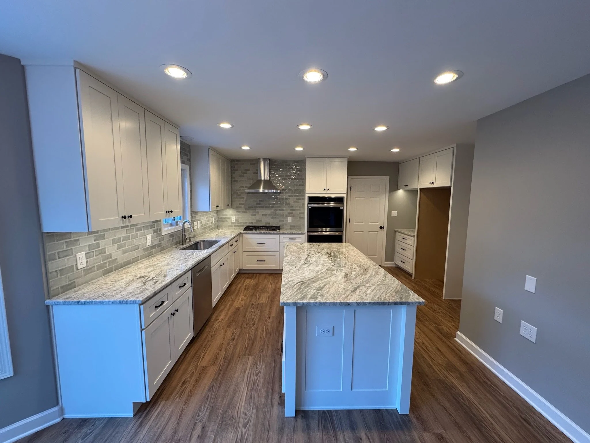A modern kitchen with white cabinets, granite countertops, a gray tiled backsplash, and recessed lighting on the ceiling. There is a central island with a matching countertop and wooden flooring throughout.