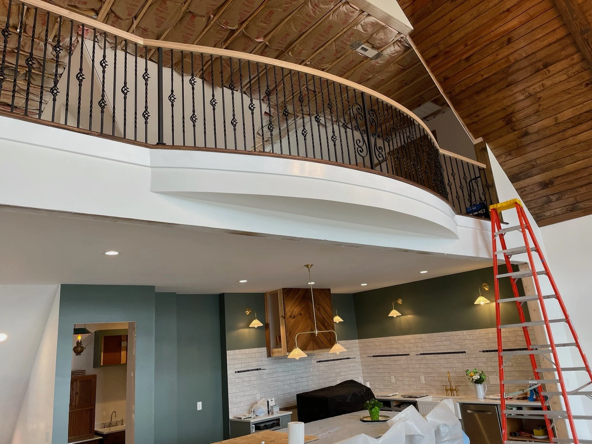 Interior view of a house renovation showing an upper balcony with black wrought iron railing, a ceiling with wood paneling, part of the kitchen with hanging pendant lights, and a red ladder nearby.