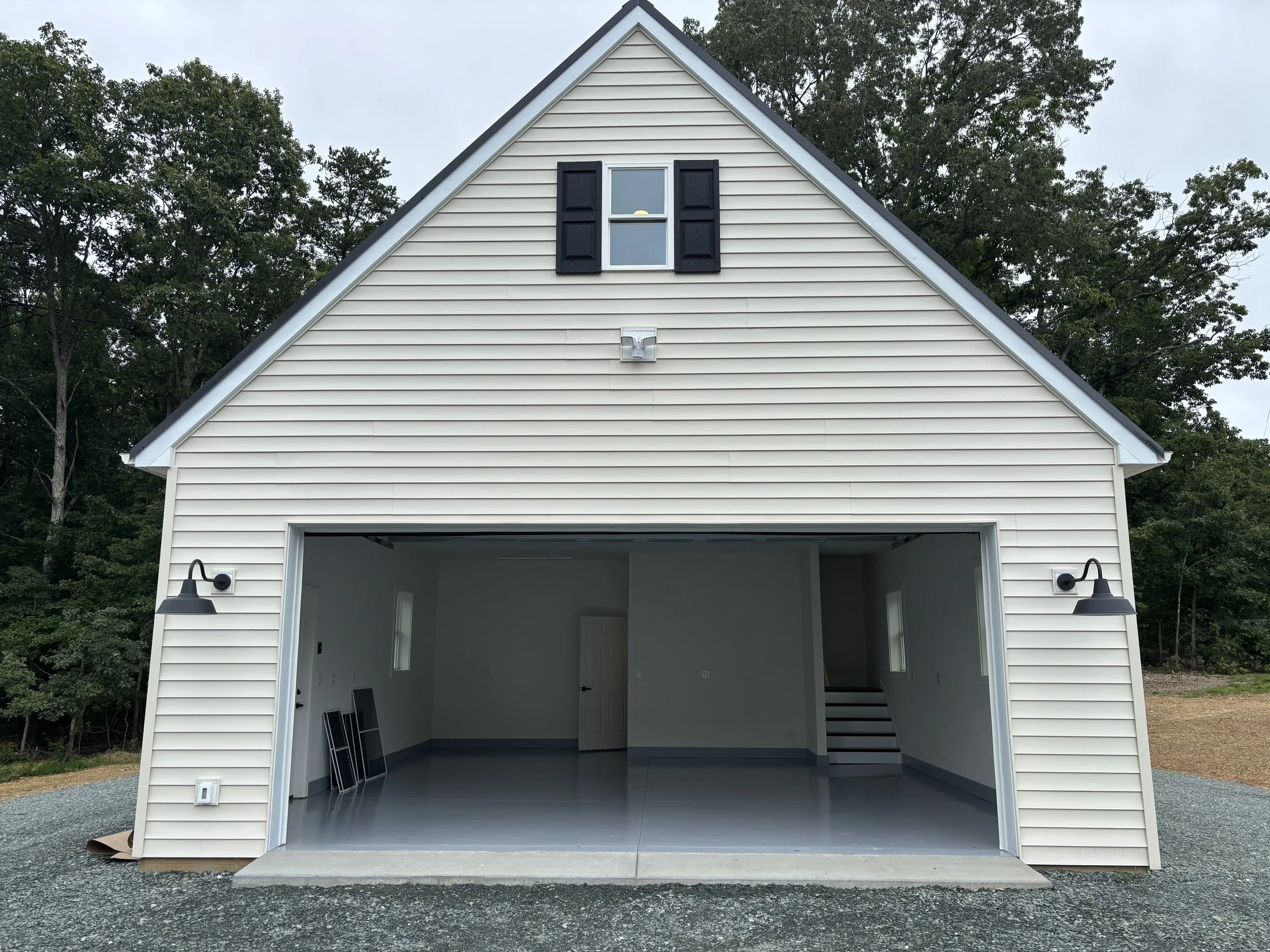 Newly constructed detached garage with open door, light-colored siding, black window shutters, and two wall sconces, set against green trees and a gravel driveway.