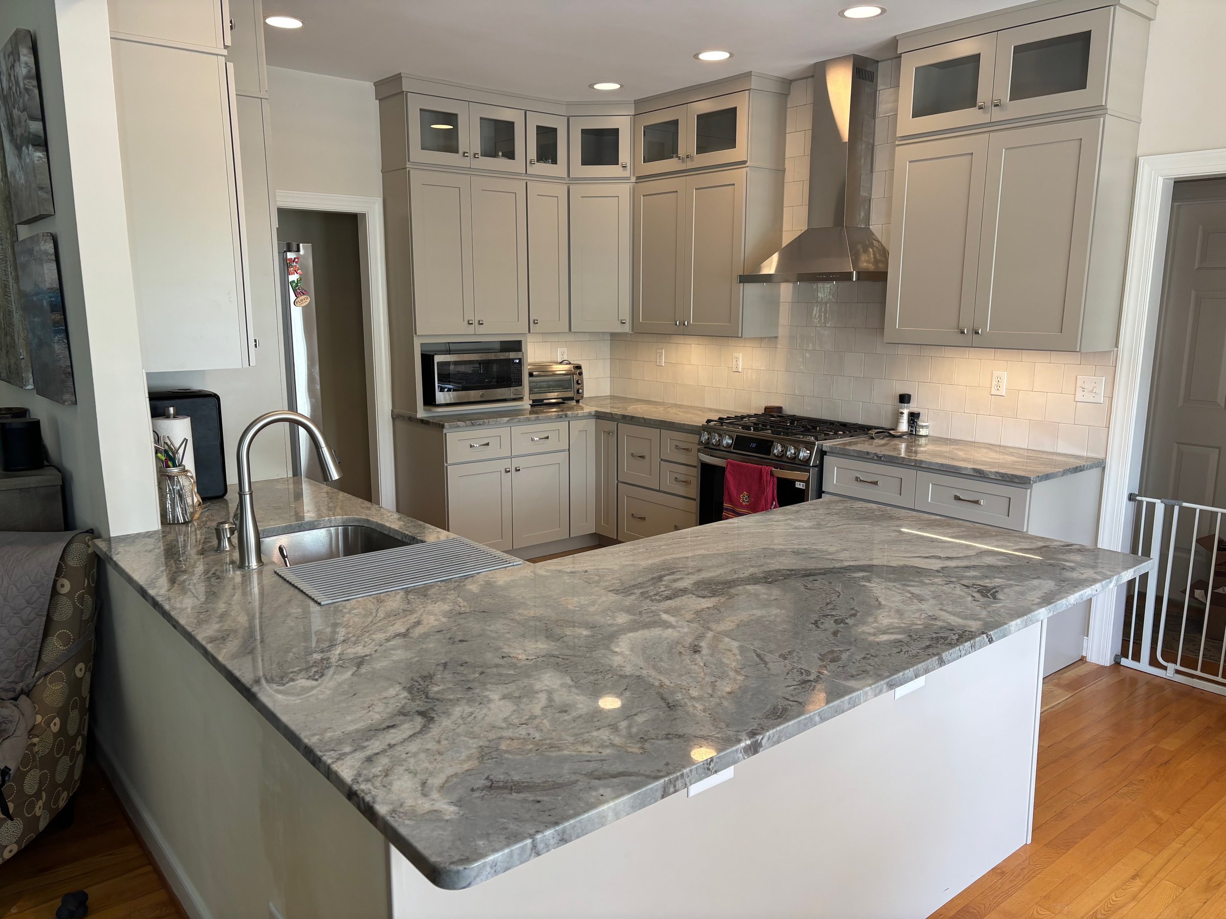 A modern kitchen with light gray cabinets, a marble countertop island, stainless steel appliances, a microwave, toaster oven, and a gas stove, with recessed ceiling lights.