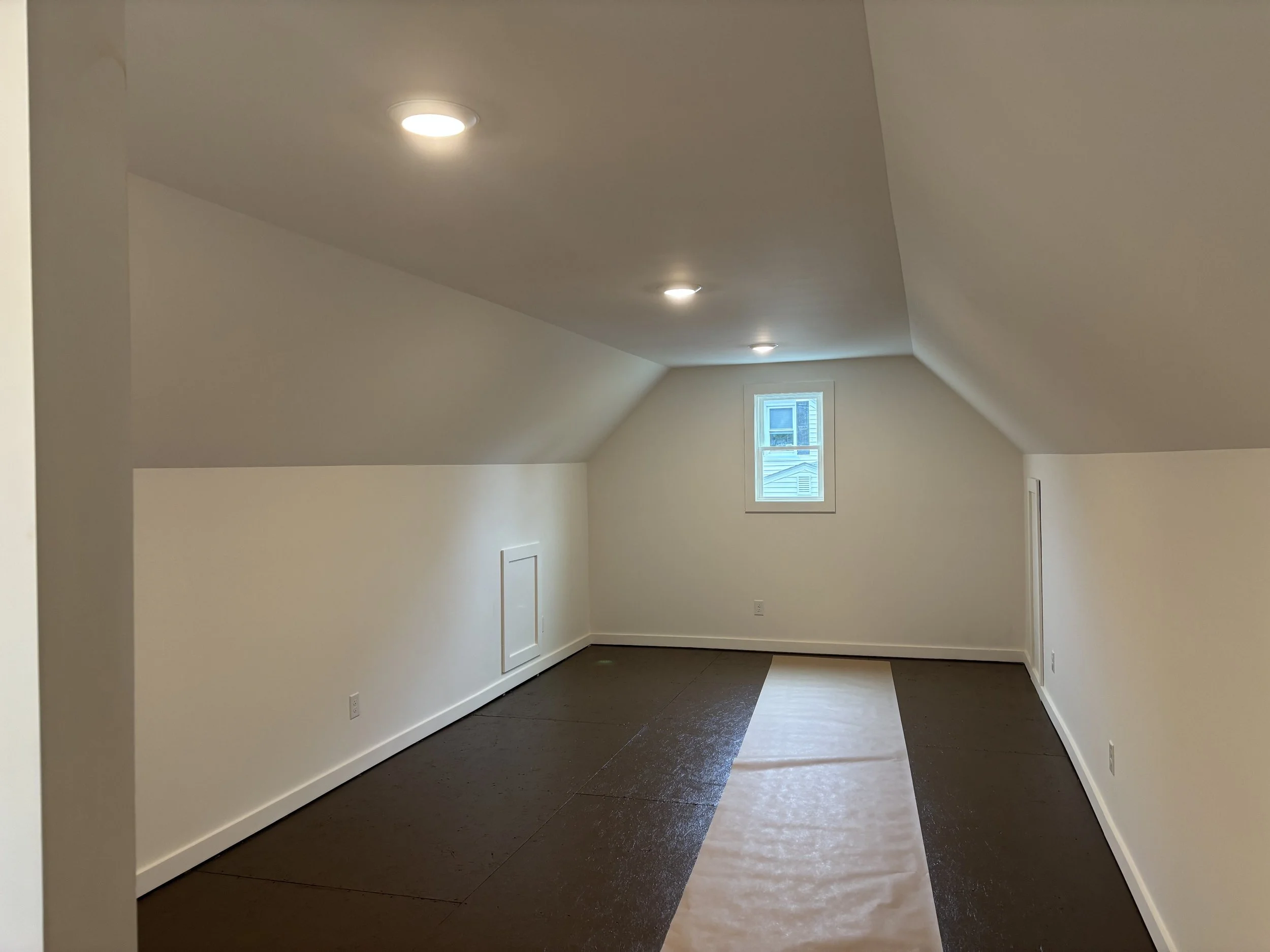 An empty attic room with sloped ceilings, a small window, and a mix of black and light brown flooring.