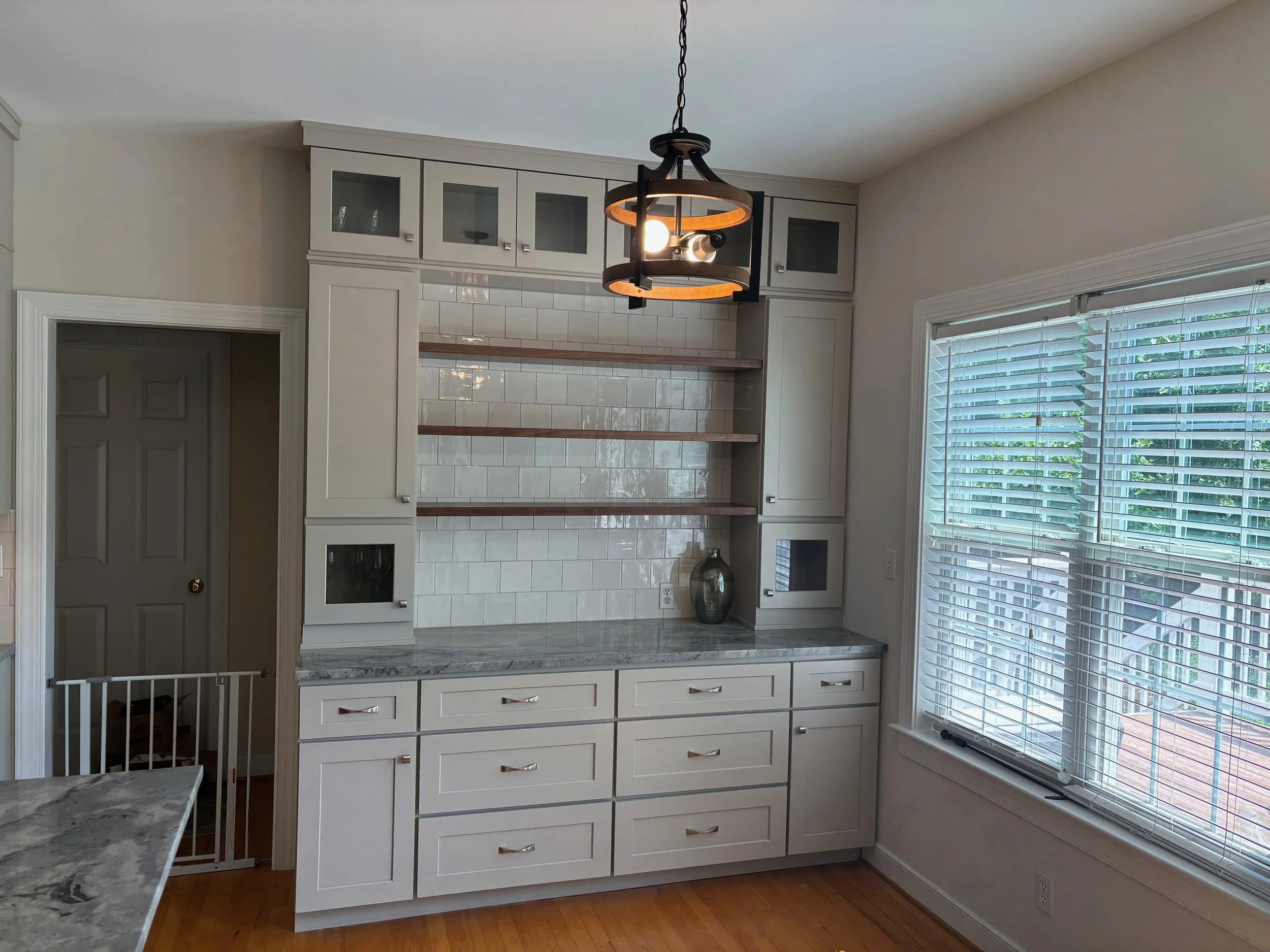 Kitchen interior with white cabinets, glass-front upper cabinets, a marble countertop, a window with venetian blinds, a chandelier, and a tiled fireplace wall.