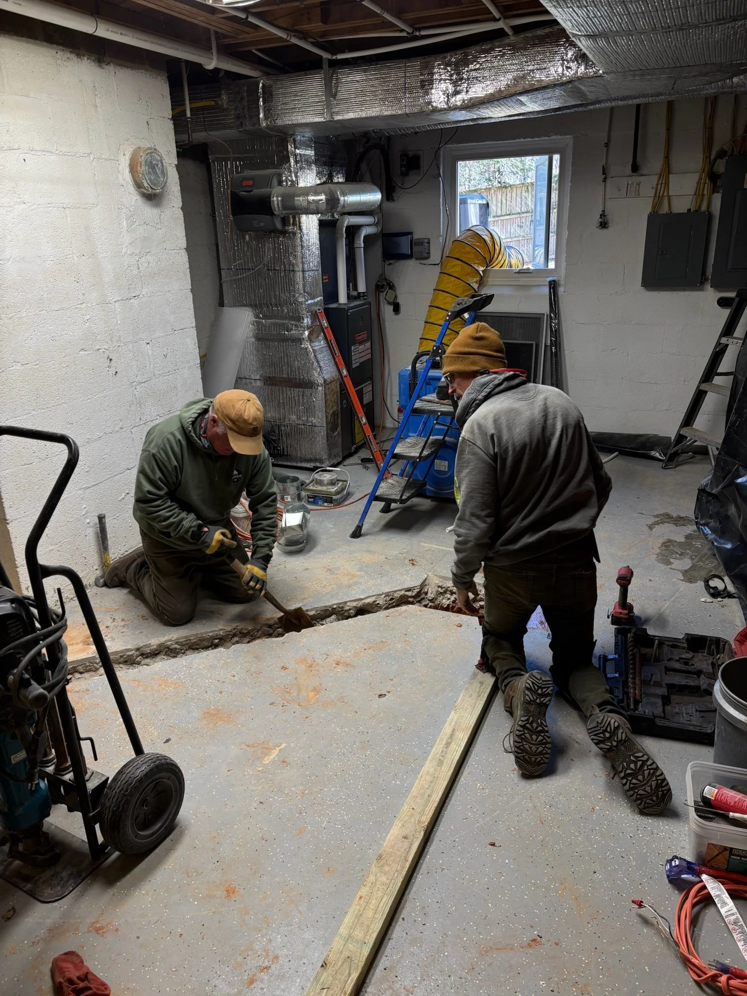 Two workers in a basement working on the floor with a long piece of wood and tools around. The basement has unfinished concrete flooring, exposed pipes, ductwork, and a small window.