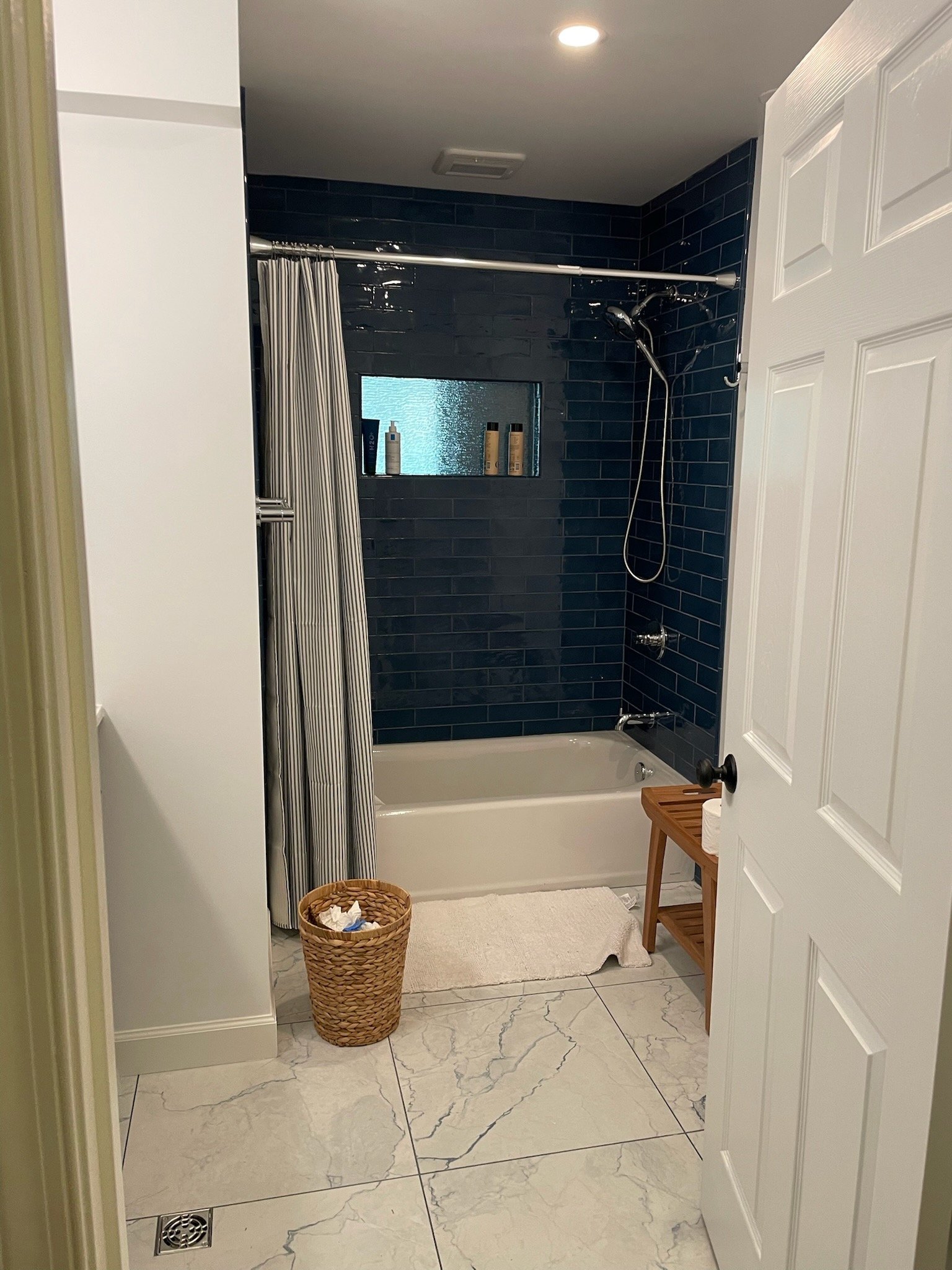 A bathroom with a shower area featuring dark blue subway tiles, and a small frosted window with bottles on the window sill. There is a white bathtub, a wicker laundry basket, a small wooden stool with toilet paper on it, and a cream-colored bath mat on the marble floor.