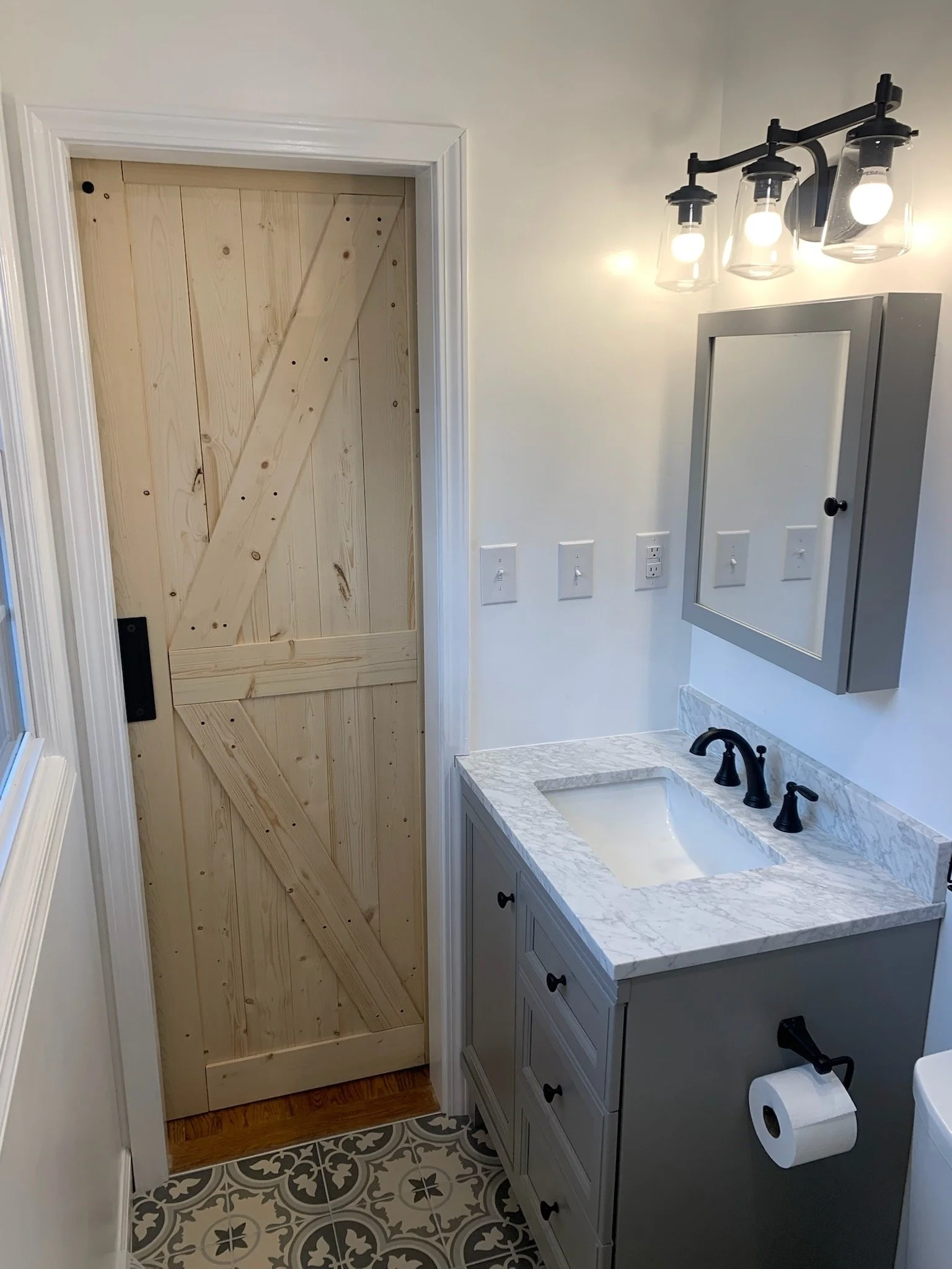 A small bathroom with a wooden door, a white vanity with a marble countertop, three black faucet handles, a mirror with a gray frame, wall-mounted light fixture with three bulbs, a toilet paper roll holder, and decorative black and white patterned floor tiles.