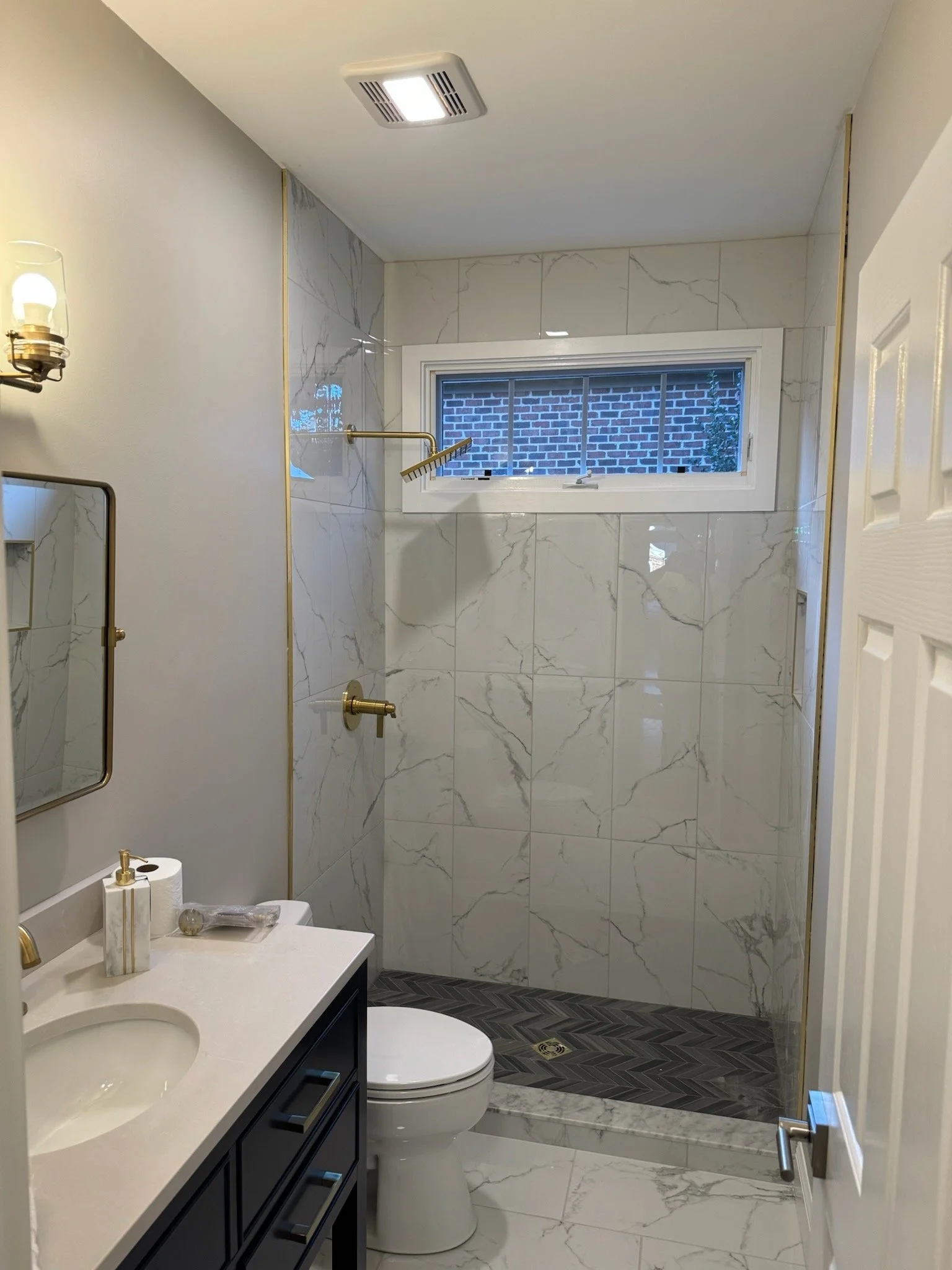 A small, modern bathroom with white marble wall tiles, a white ceiling, and a small horizontal window. It features a black vanity with a white countertop and a round sink, a toilet, and a shower with a gold showerhead and fixtures. There are gold accents on the wall and a mirror is partially visible.