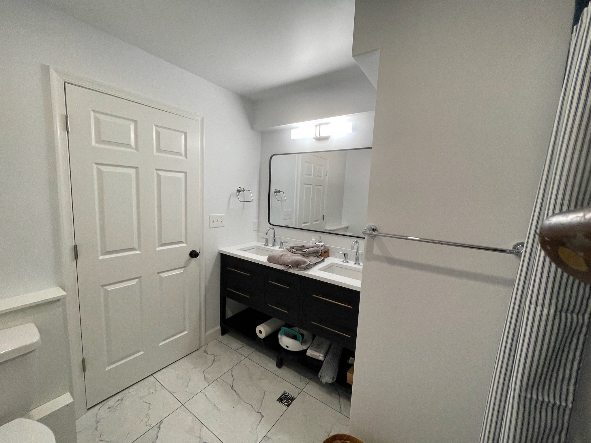 A bathroom with white walls, a white door, and a large mirror above a two-sink vanity with a white countertop and black cabinet. Towels are on the counter, and a towel rack is on the wall. The floor has white marble tiles with gray veins, and there's a window near the ceiling with light streaming in.