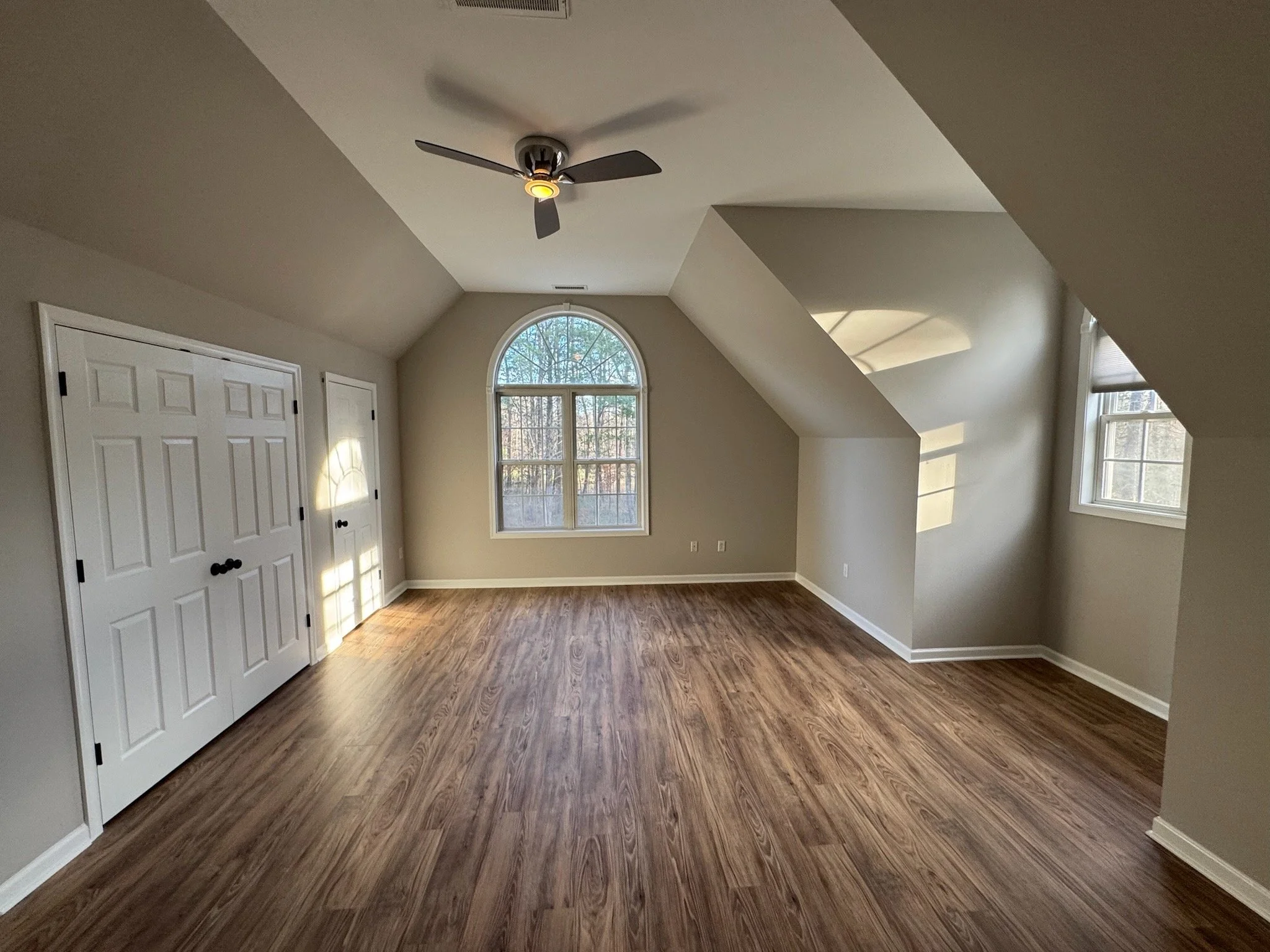 A spacious room with large arched window, smaller rectangular windows, two white closet doors, hardwood flooring, light gray walls, and a ceiling fan.