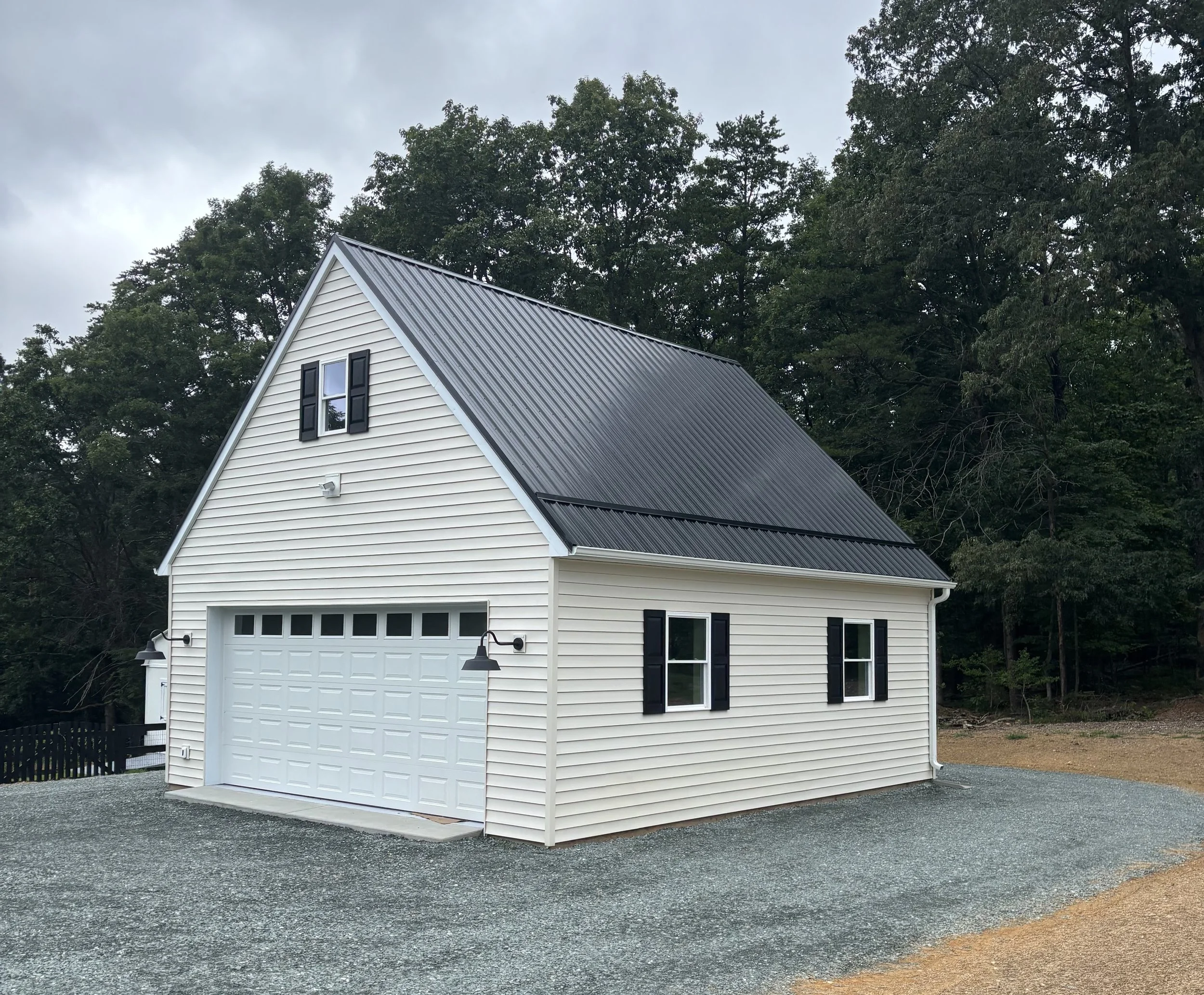 A white house with black shutters and a metal roof, set against a background of tall green trees and cloudy sky.