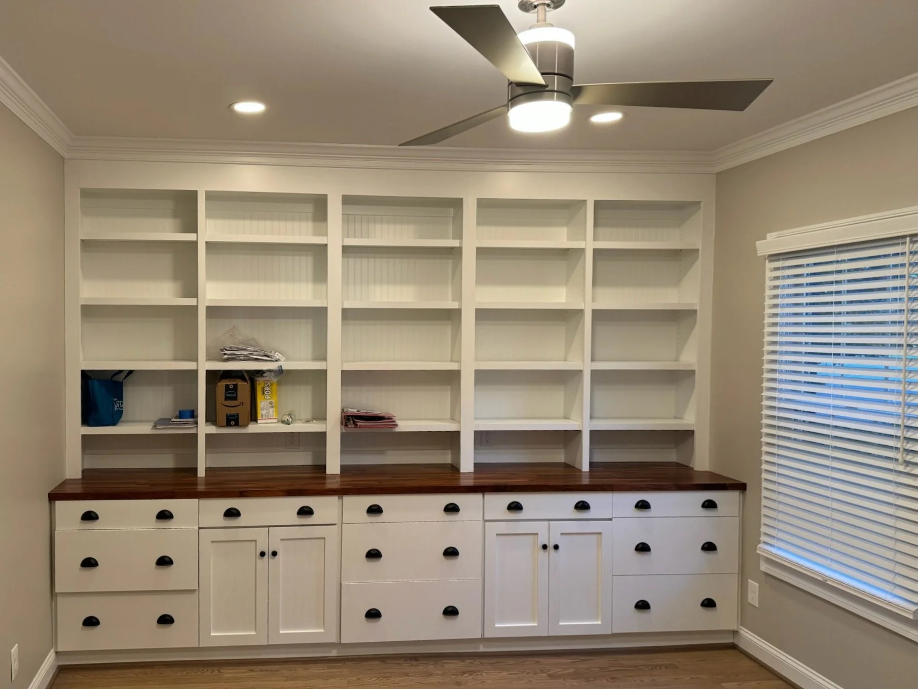 Empty white built-in bookshelf with six shelves and a lower cabinet with black round knobs, located in a room with beige walls, crown molding, a ceiling fan, and a window with white blinds.