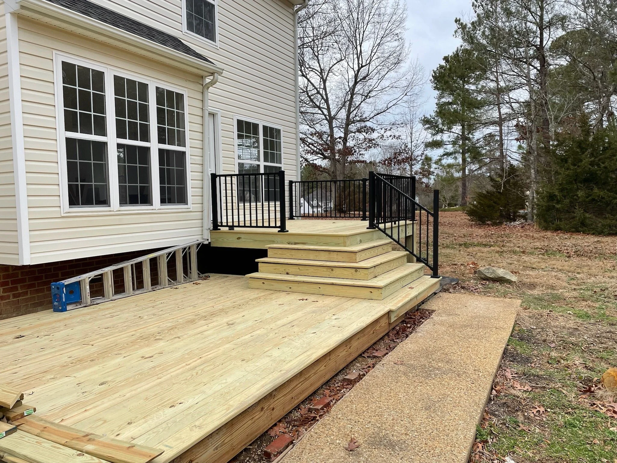 A newly built wooden deck attached to the back of a house with white siding, at an early stage of construction with stairs and black metal railing, surrounded by a yard with trees and fallen leaves.