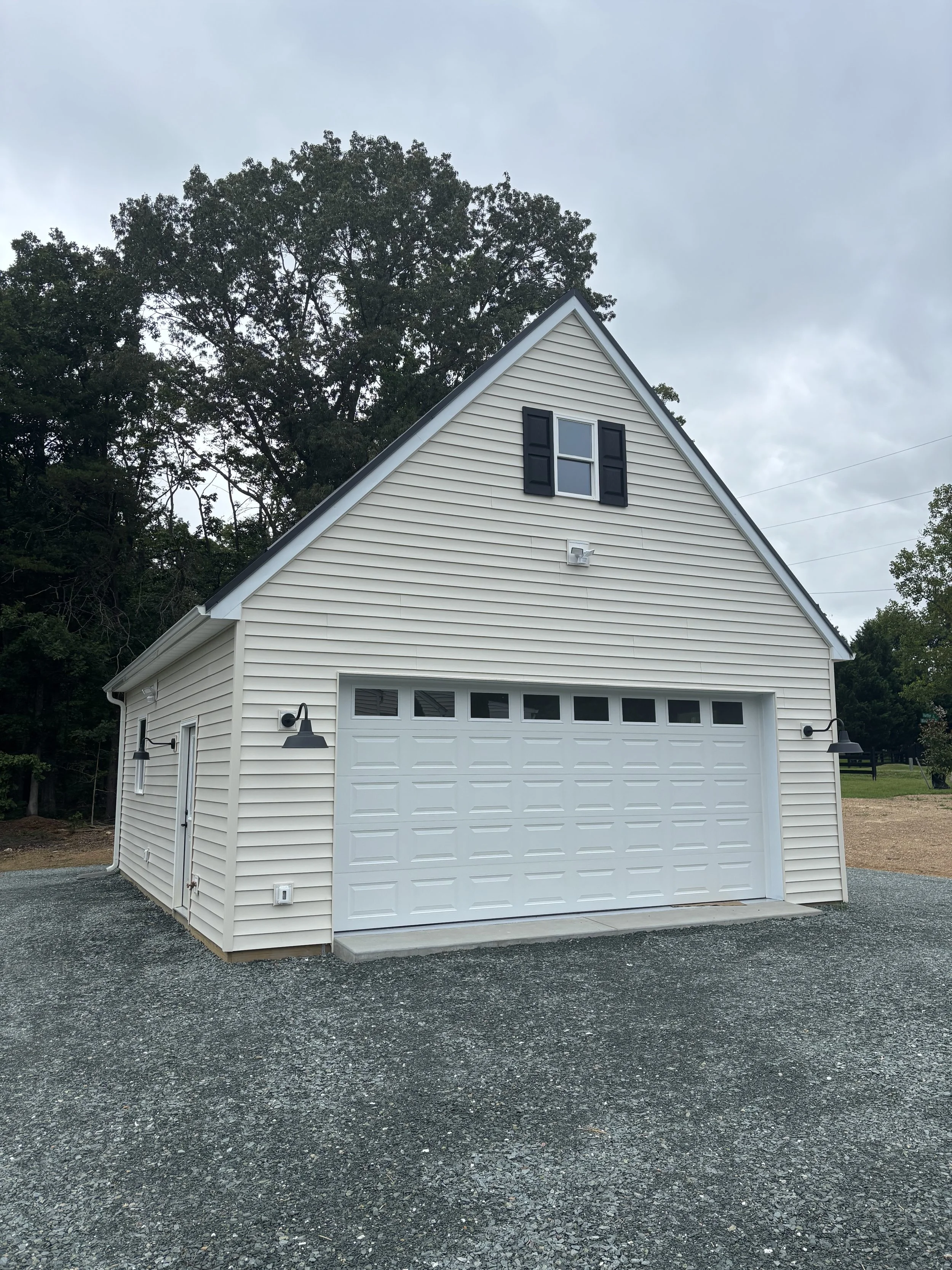A white garage with a pitched roof and black shutters, surrounded by trees, with two black wall-mounted lamps, gravel driveway, and a cloudy sky overhead.