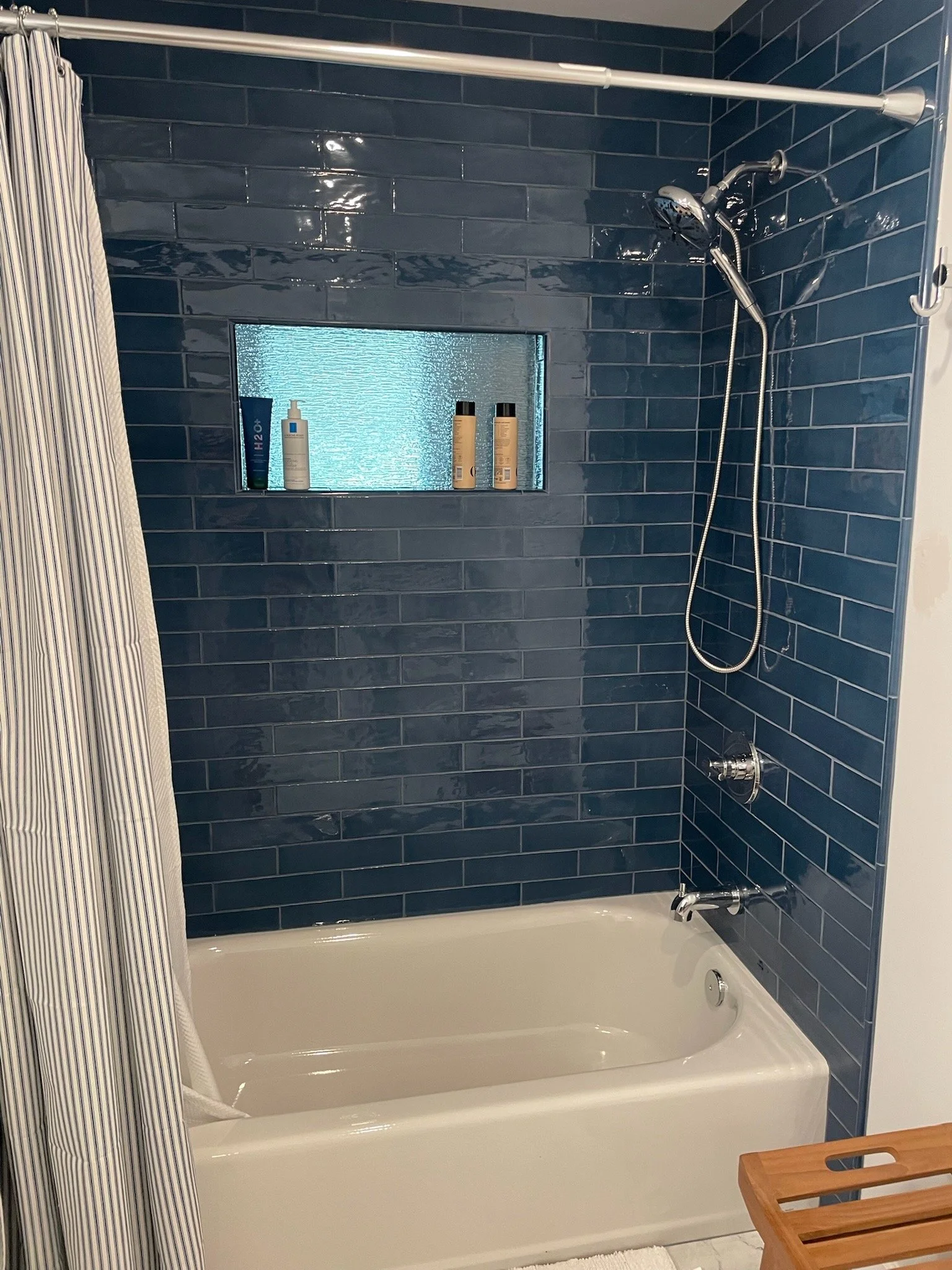 A bathroom with a dark blue tiled wall, a bathtub, a shower head with a flexible hose, bottles of shower products on a small window shelf, a striped shower curtain, and a wooden bath tray.