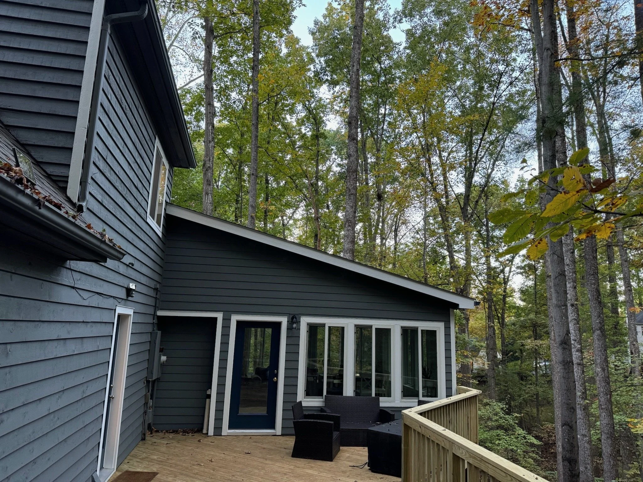 A wooden deck attached to a house with dark gray siding, featuring a black patio table and chairs, overlooking a forest of tall trees with green leaves.