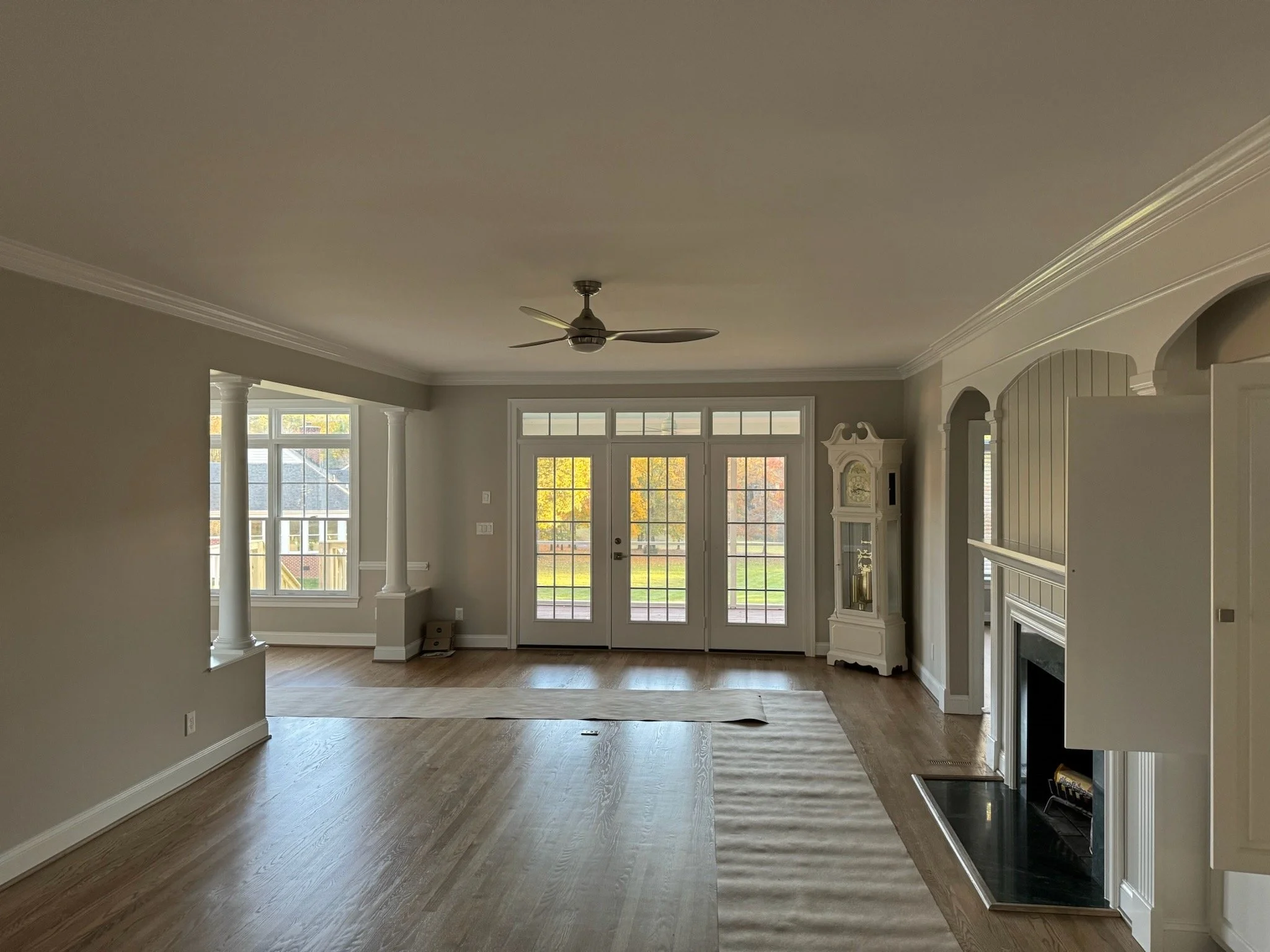 Interior view of a spacious living room with large windows, a ceiling fan, hardwood floors, and built-in shelves around a fireplace.