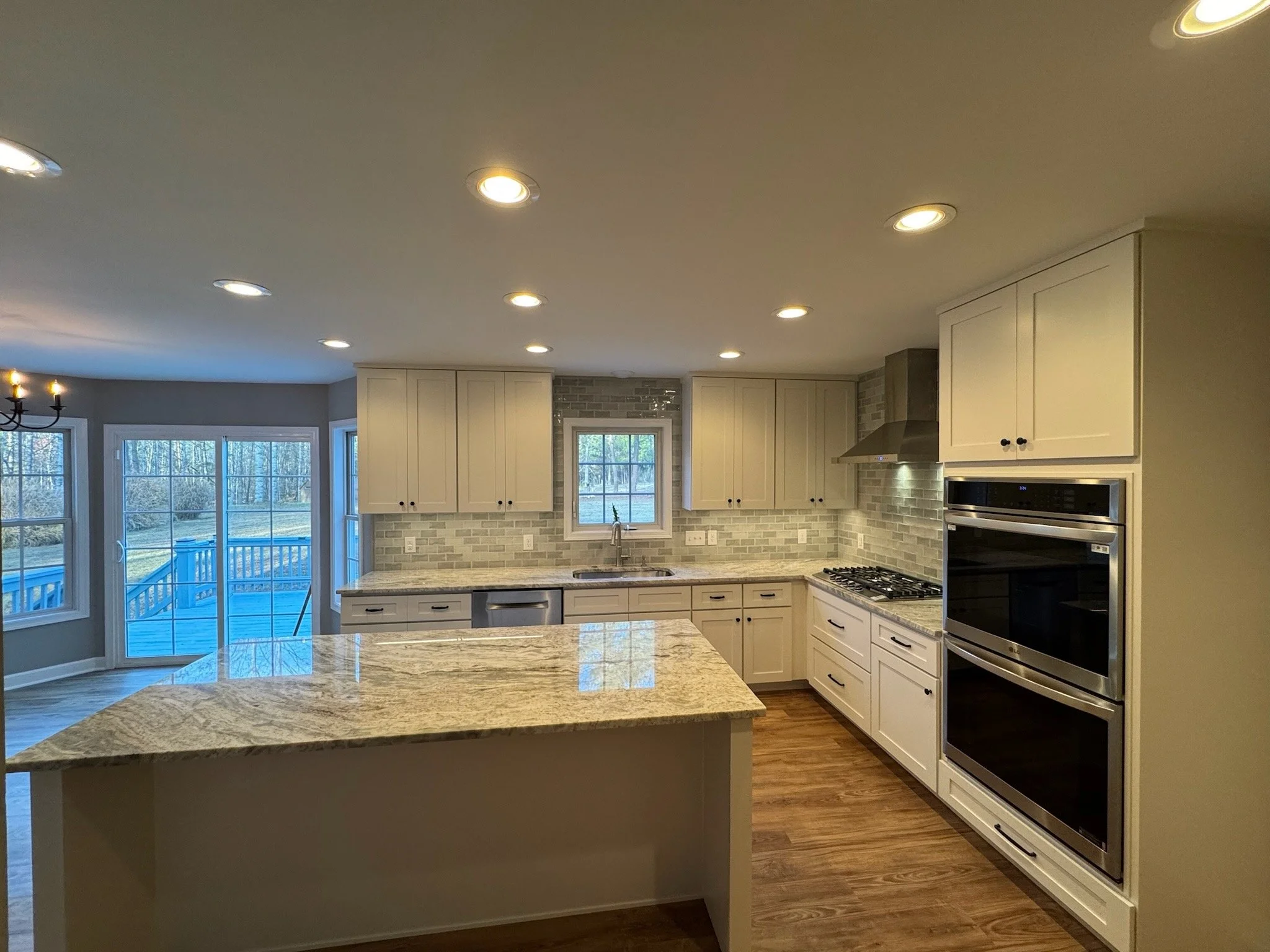 Modern kitchen with white cabinets, a marble countertop island, stainless steel oven, brick backsplash, and large windows overlooking a backyard deck with trees.