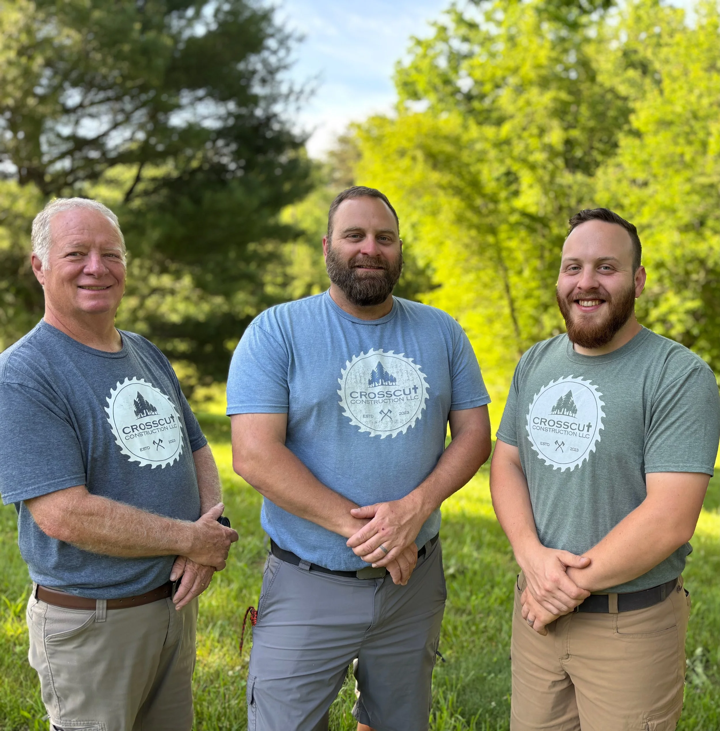 Three men standing outdoors in a park with green trees, smiling and wearing casual shirts with a logo for Crosscut Construction LLC.