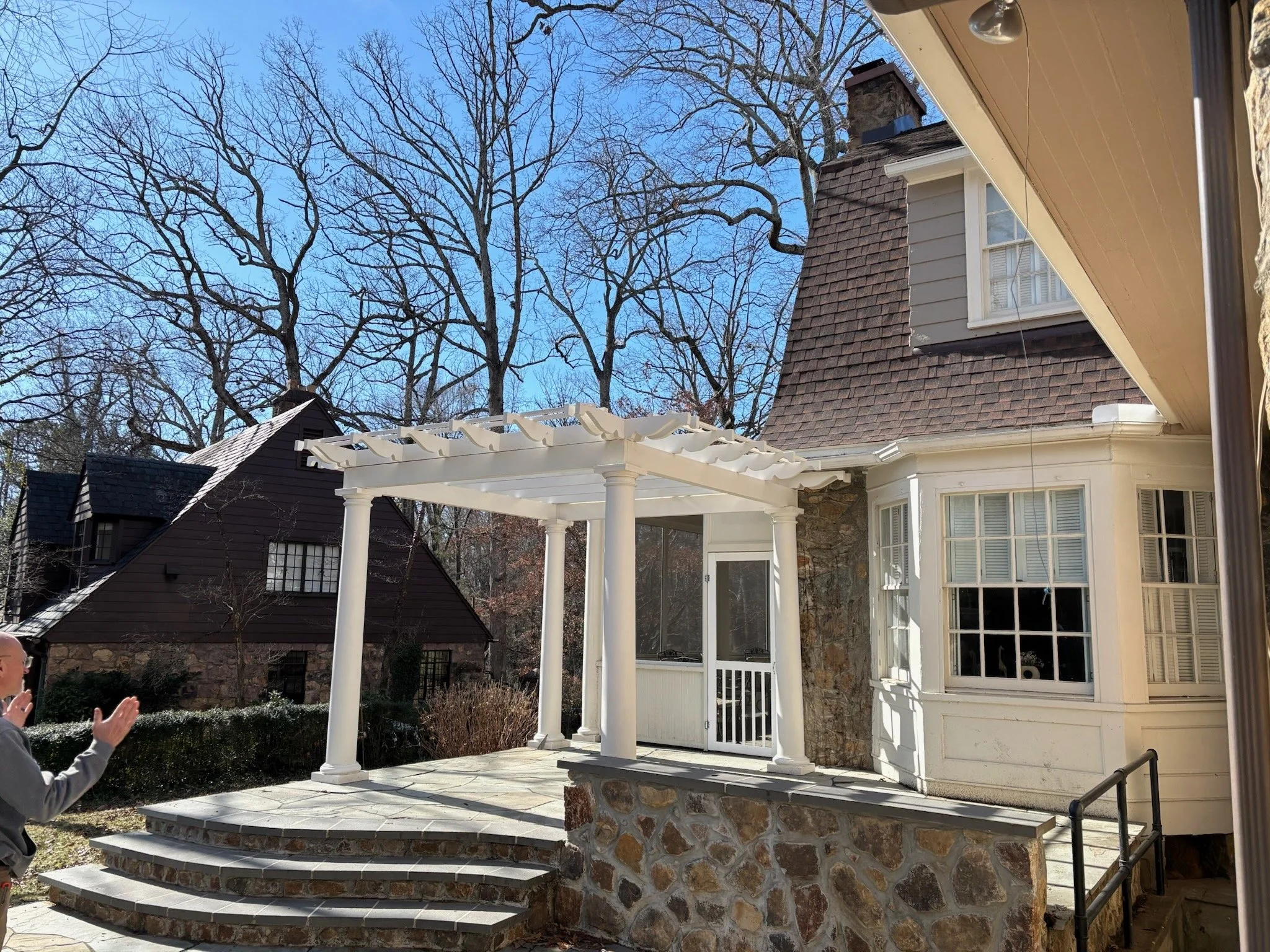 A backyard patio with a white pergola, stone stairs, and a house with beige siding and a stone chimney. Bare trees are visible in the background under a clear blue sky.