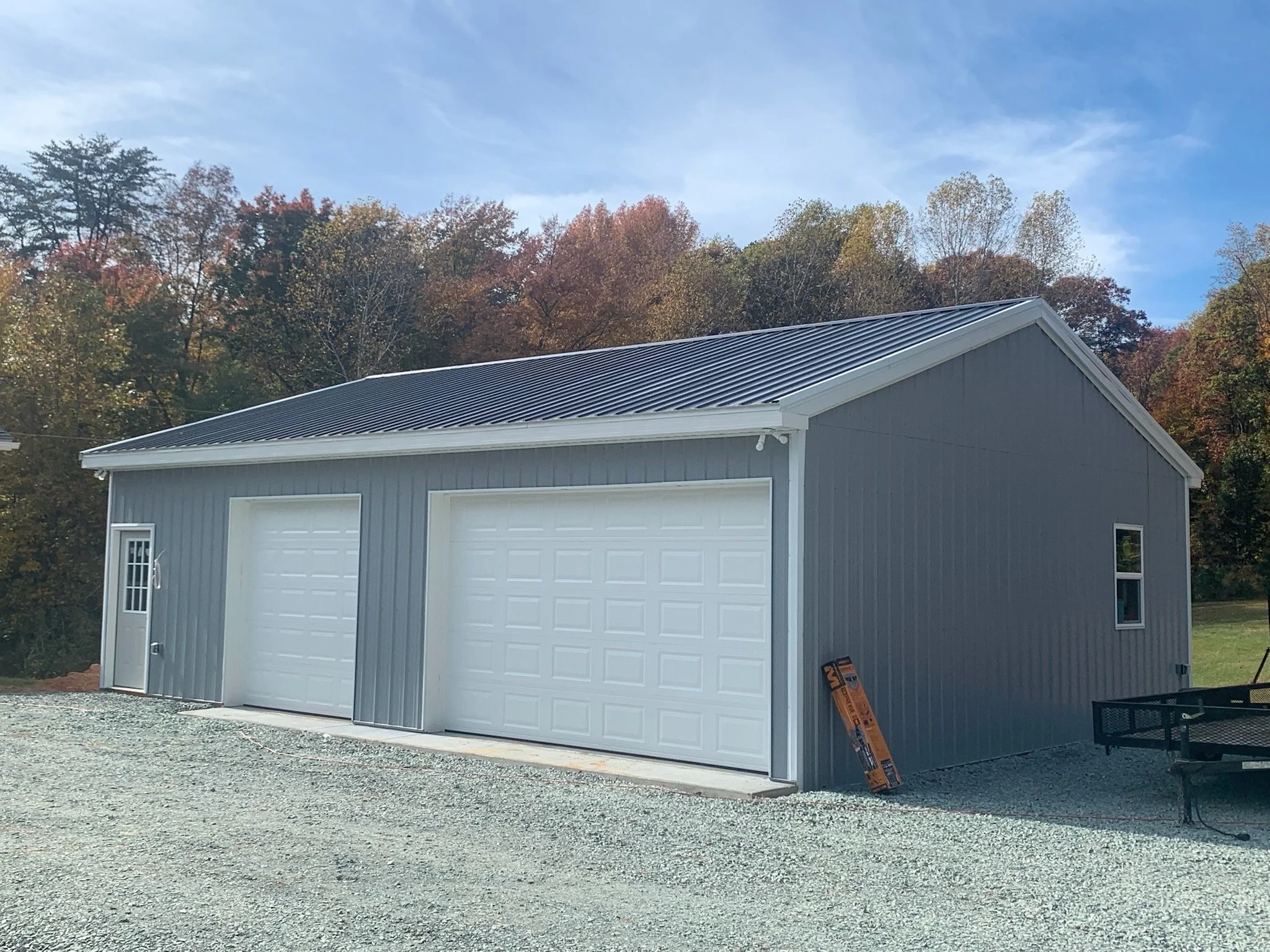 A gray garage with white trim and a closed garage door, with a small window on the side, a gravel driveway, and a background of autumn-colored trees and a blue sky with some clouds.