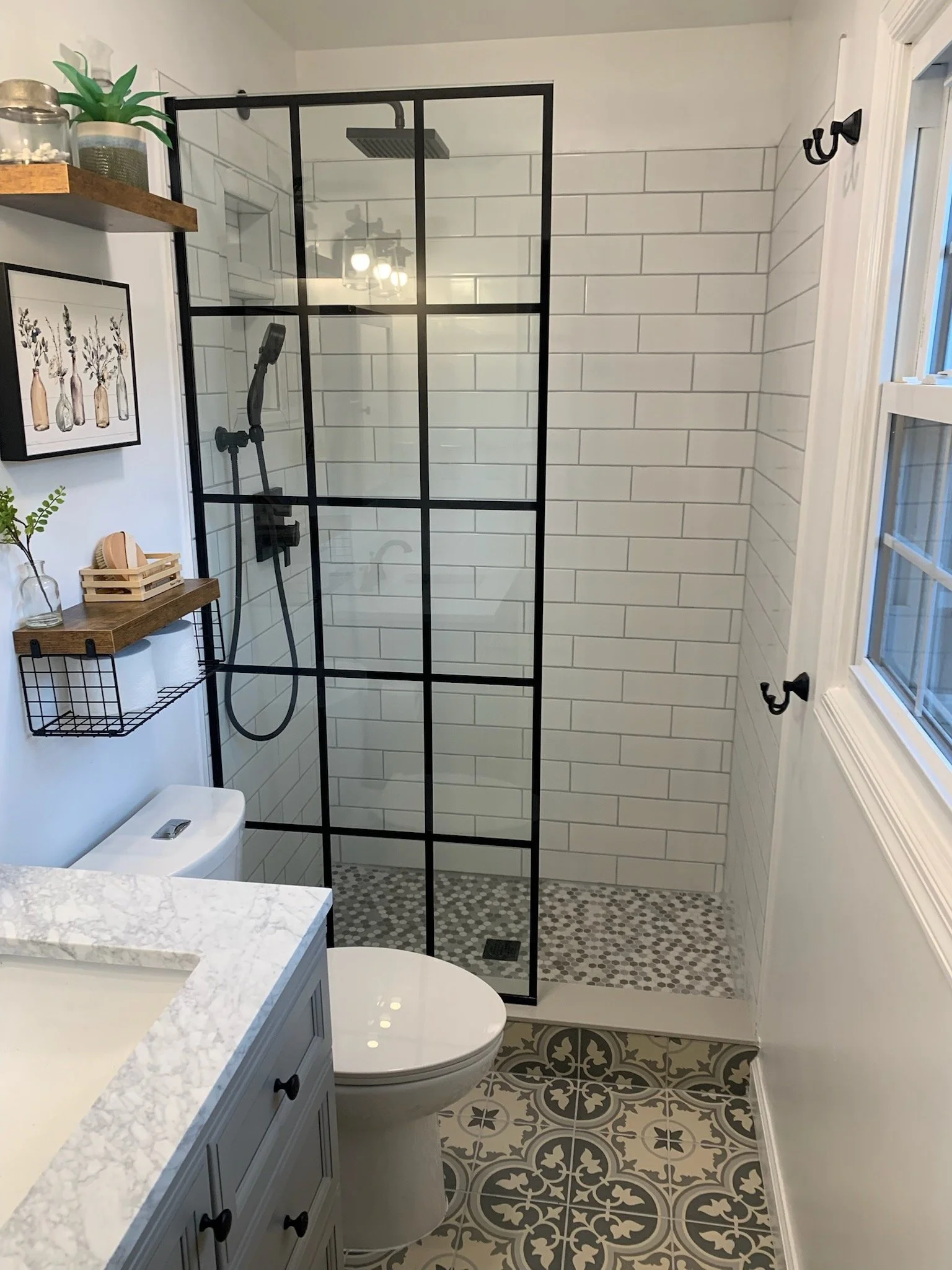 A modern bathroom featuring a walk-in shower with clear glass black metal frame, white subway tile walls, patterned floor tiles, a white toilet, a blue vanity with a white marble countertop, decorative plants, wall art, and black hooks on the wall.