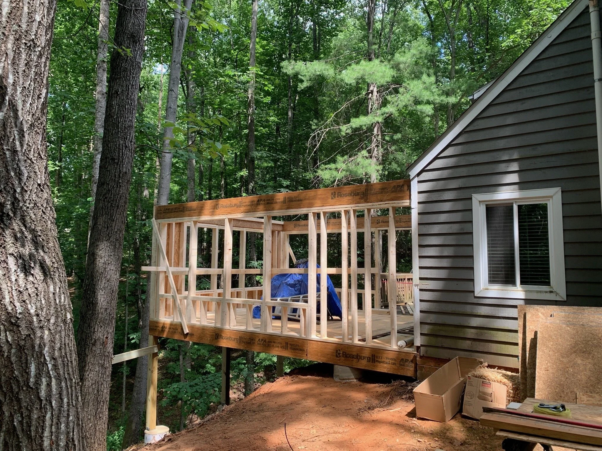 A house under construction among trees, with a partially built wooden deck or porch attached to the house. Construction materials and tools are visible on the ground and nearby.