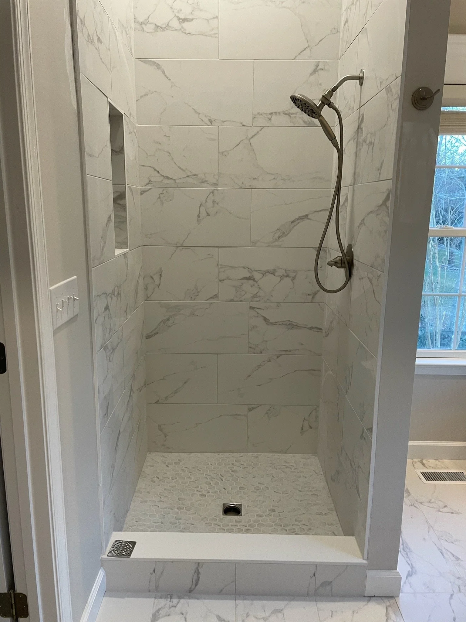 Empty walk-in shower with marble wall tiles, pebble floor, glass door, and window showing outside trees.