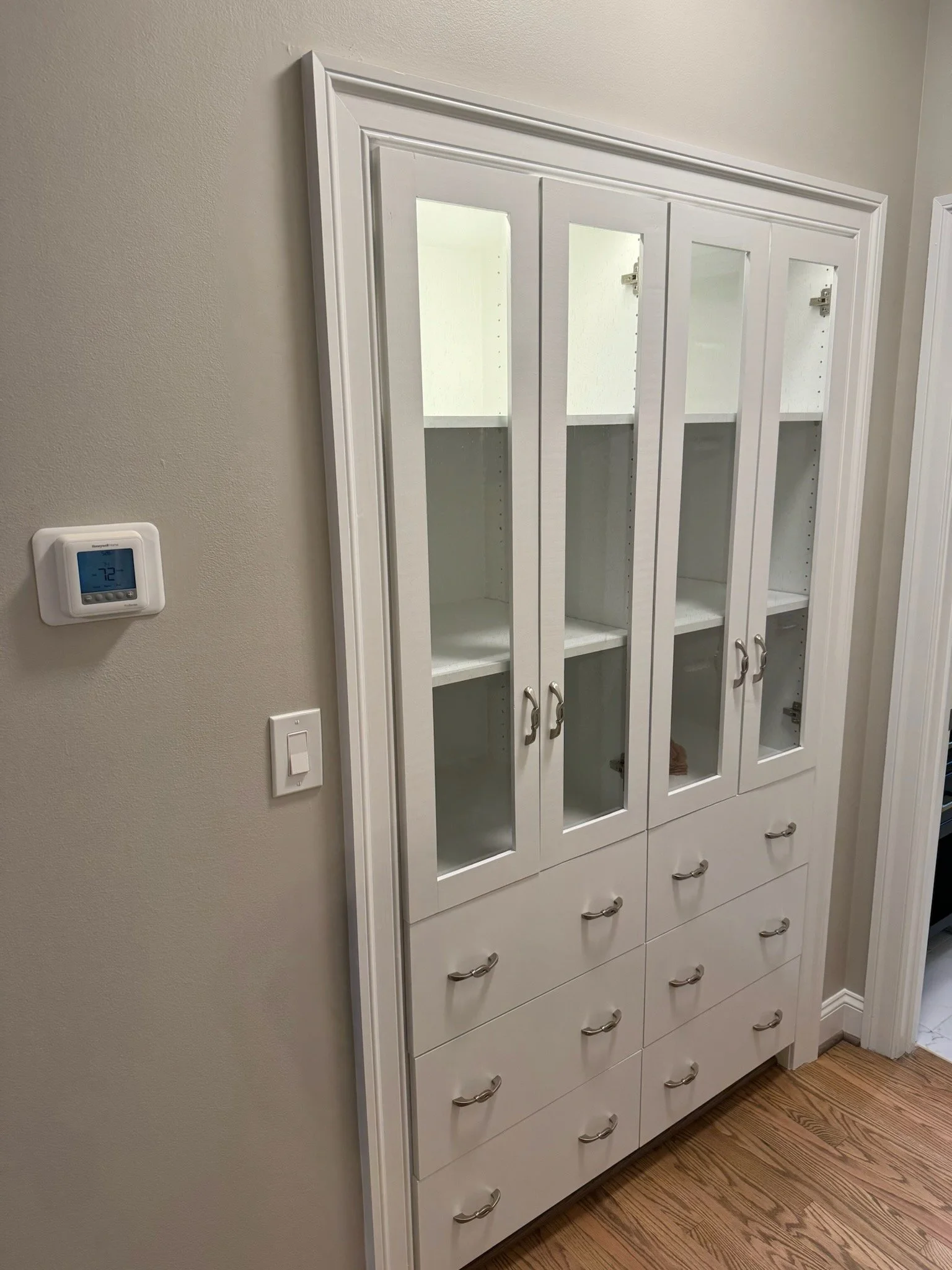 White display cabinet with glass doors and drawers below, installed against a beige wall, with a thermostat and light switch nearby, on a hardwood floor.