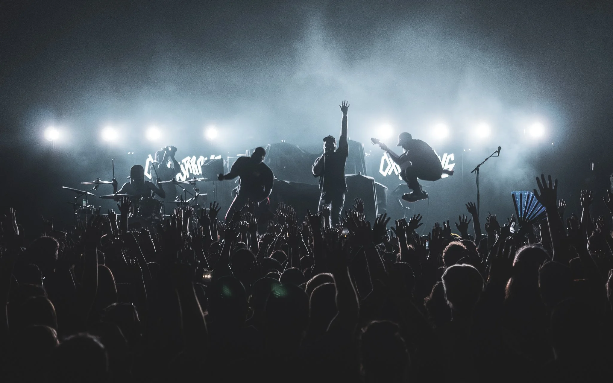 Silhouettes of band members performing on stage, with an enthusiastic crowd in front, raised hands, and bright stage lights in the background.